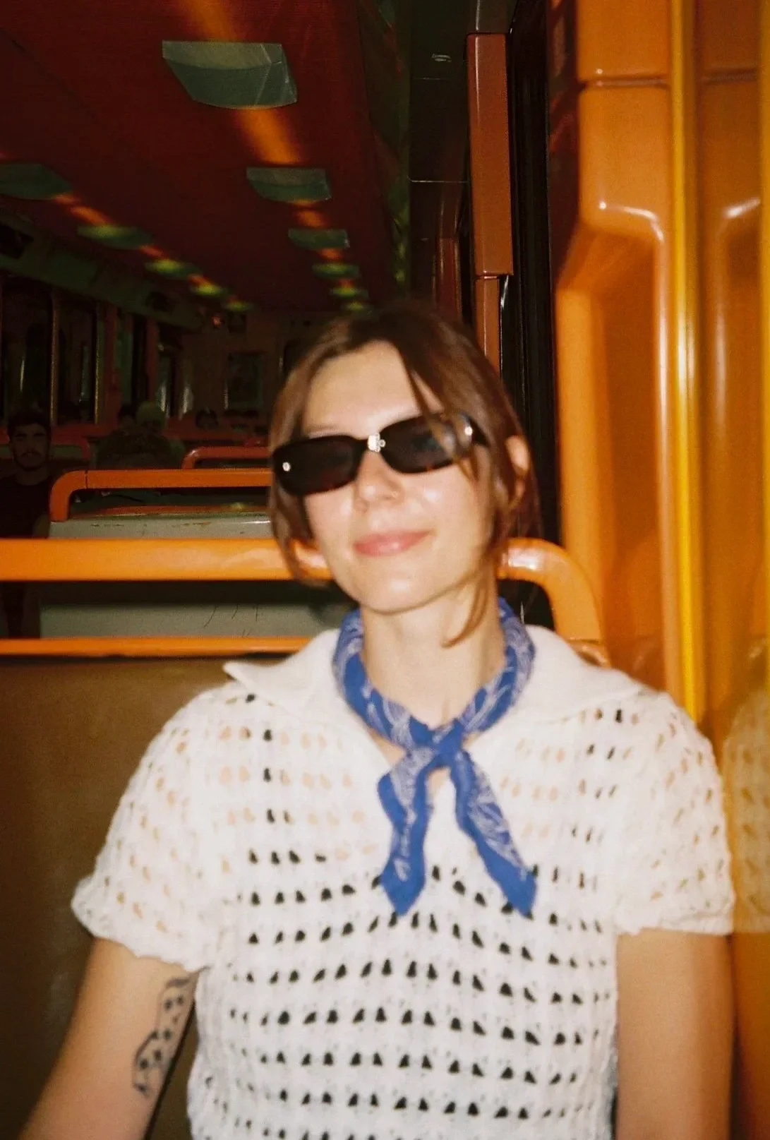 A woman with short brown hair wearing black sunglasses, a white crochet top, and a blue scarf around her neck, sitting inside a colorful bus or train.