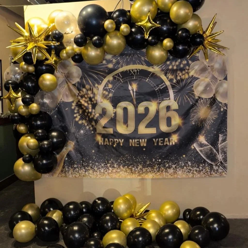 Decorative New Year 2026 celebration setup with black, gold, and clear balloons arranged around a backdrop featuring fireworks, a clock about to strike midnight, and the text '2026 Happy New Year'.