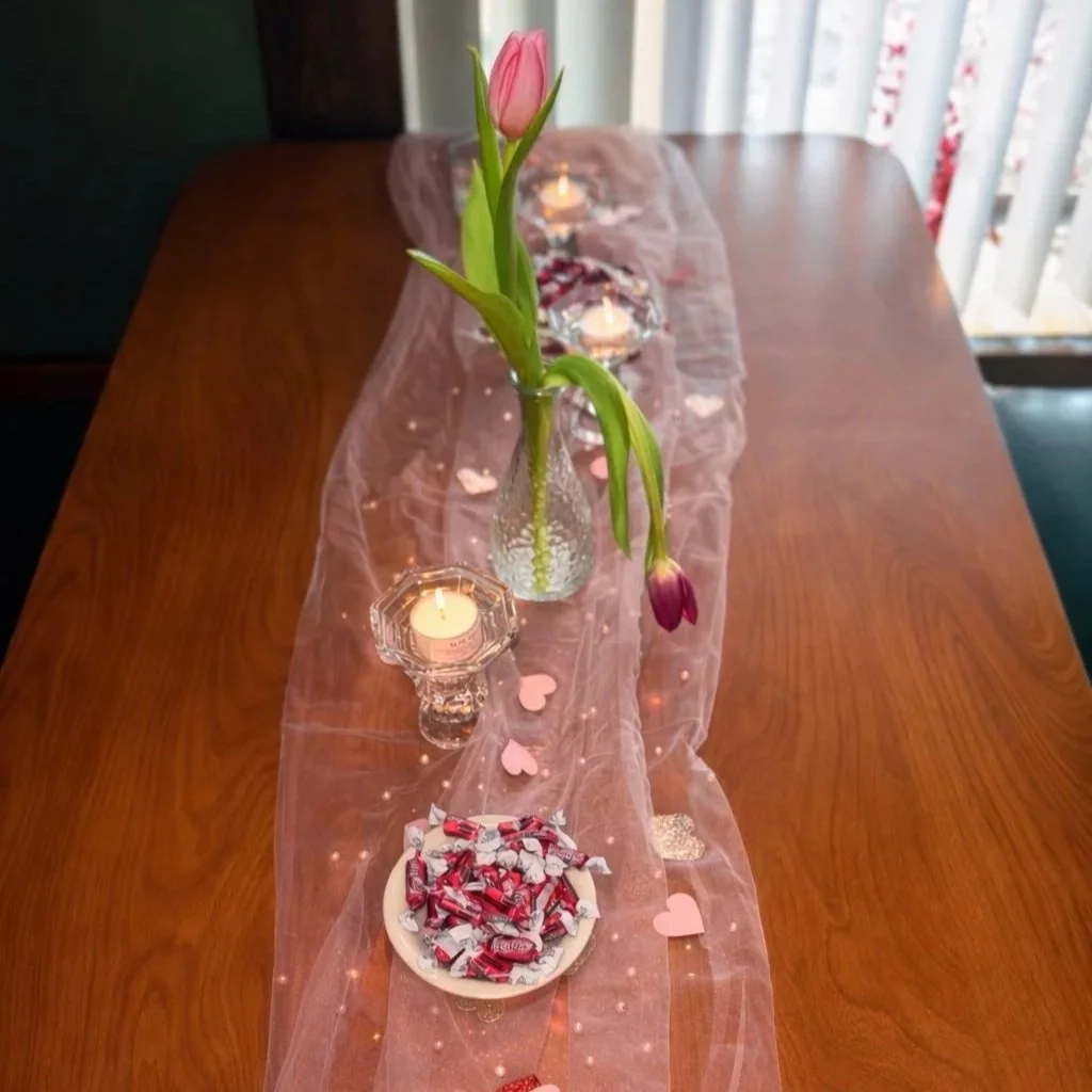 Table decorated with a pink sheer table runner, pink tulips in a small vase, candles, heart-shaped decorations, and a bowl of wrapped candies.