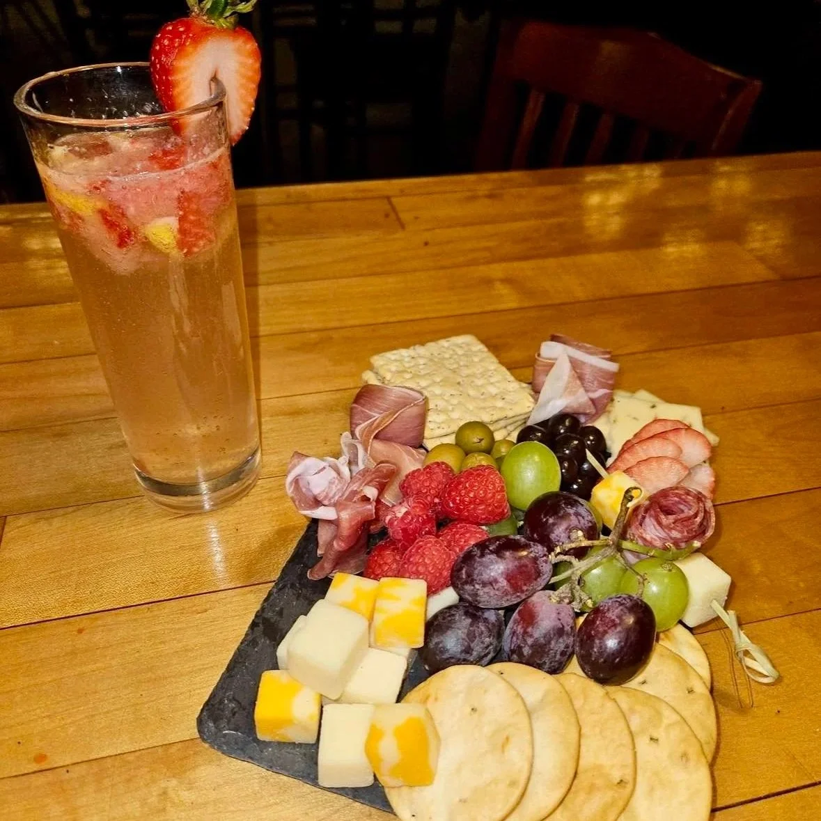 A cheese and fruit platter with grapes, raspberries, strawberries, sliced cheese, and meats, alongside a tall glass of sparkling drink with strawberries, on a wooden table.