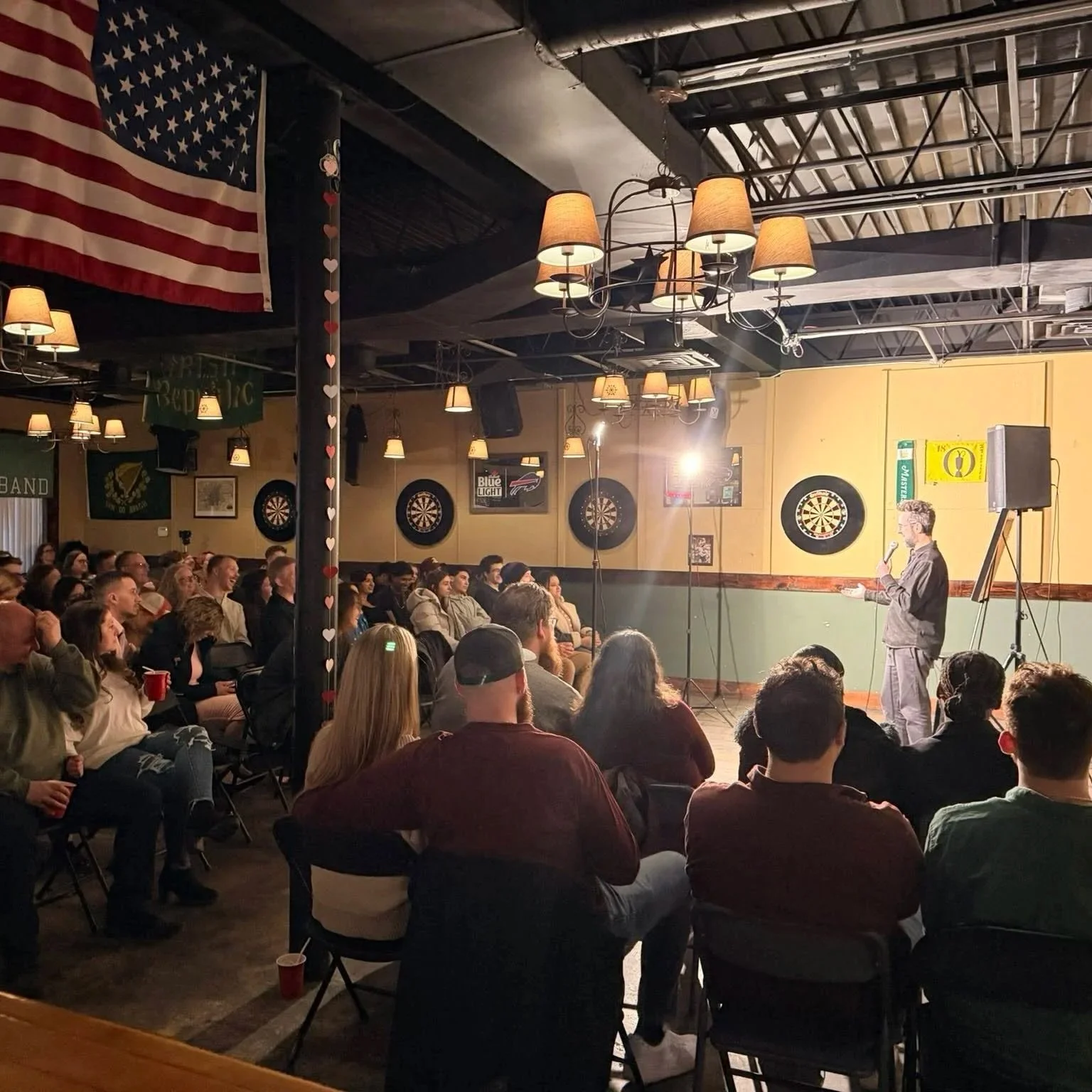 A man with glasses stands on a stage with a microphone, speaking to an audience in a dimly lit bar or comedy club. The audience is seated facing the stage, and the room is decorated with chandeliers, dartboards, and an American flag. The ceiling has exposed pipes and ductwork.