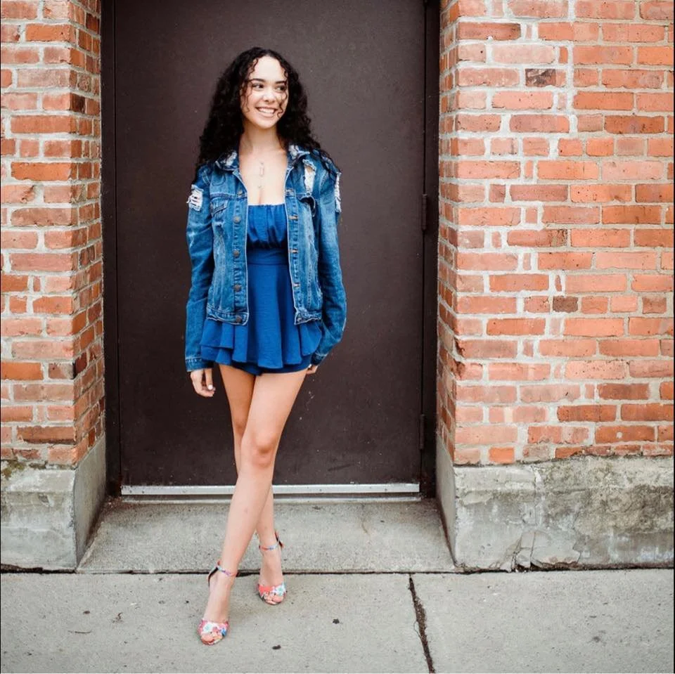 A young woman with curly hair smiling, standing in front of a brick wall and black door, wearing a blue dress, denim jacket, and floral heels.