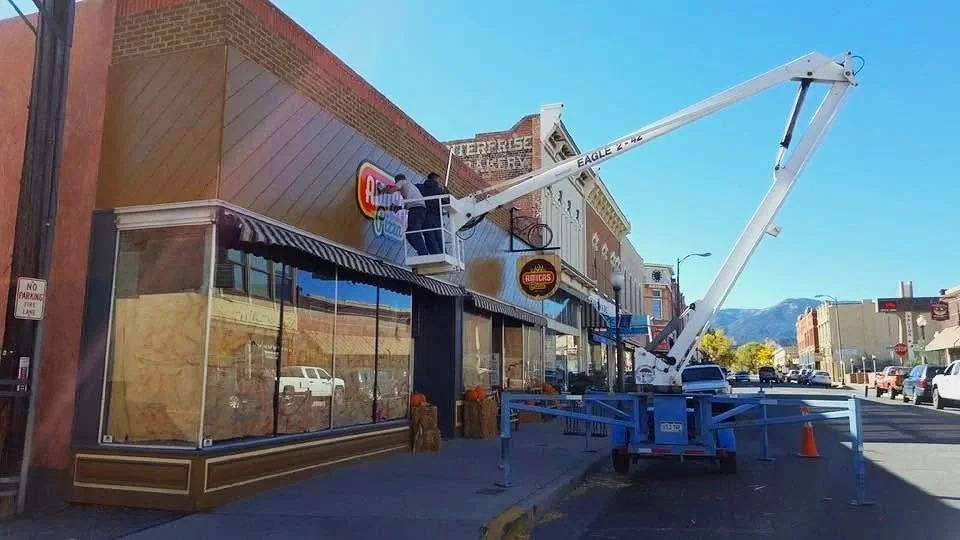 A worker on a cherry picker cleaning the exterior window of a restaurant or cafe on a city street. The scene shows a clear sky, parked cars, and mountain range in the background.