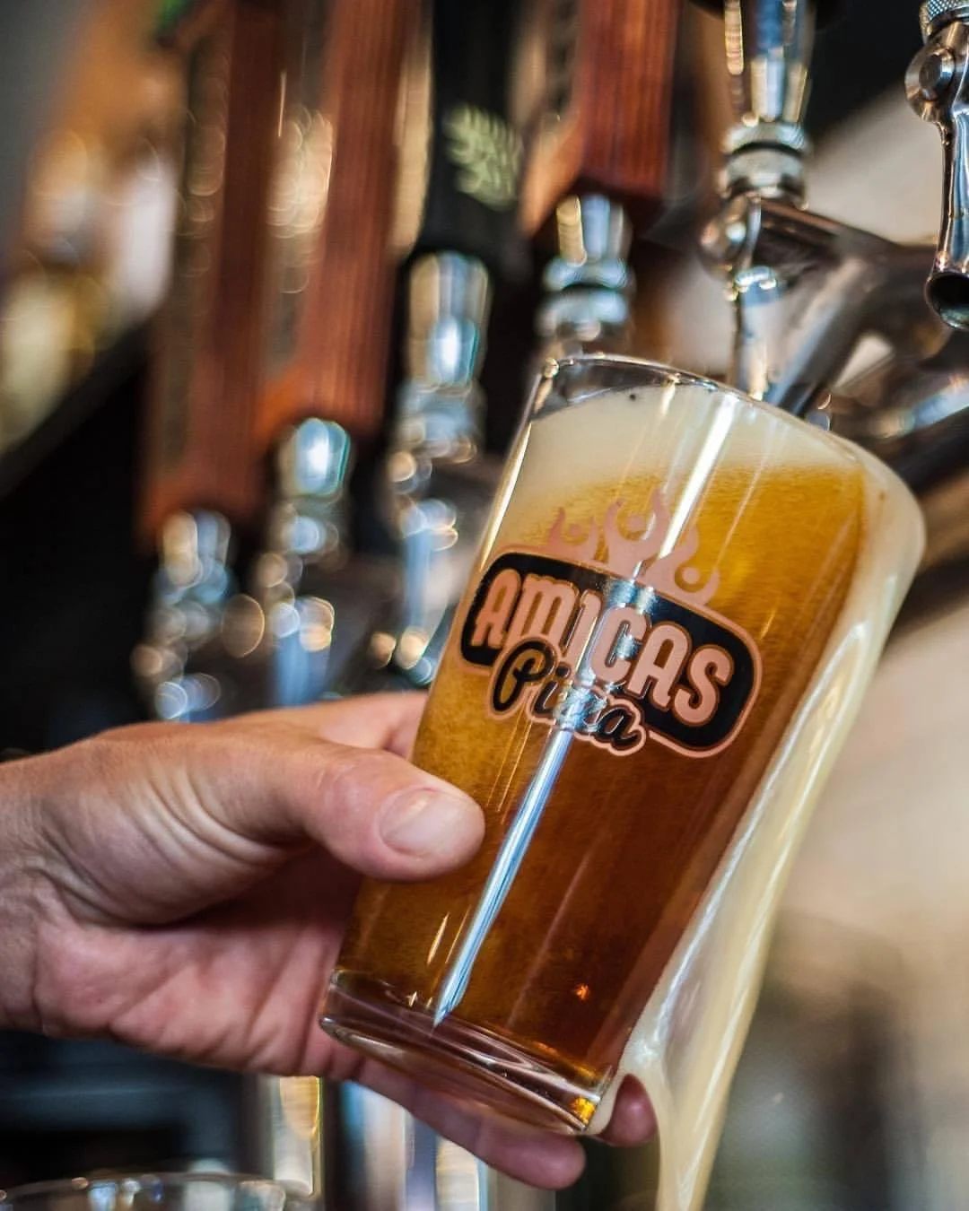 A hand holding a glass of amber beer with a logo that says 'Americanos' on it, in front of a row of beer taps.