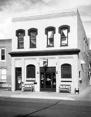 Black and white photo of a two-story brick building with arched windows and a central entrance with an awning, benches outside, and a sidewalk in front.