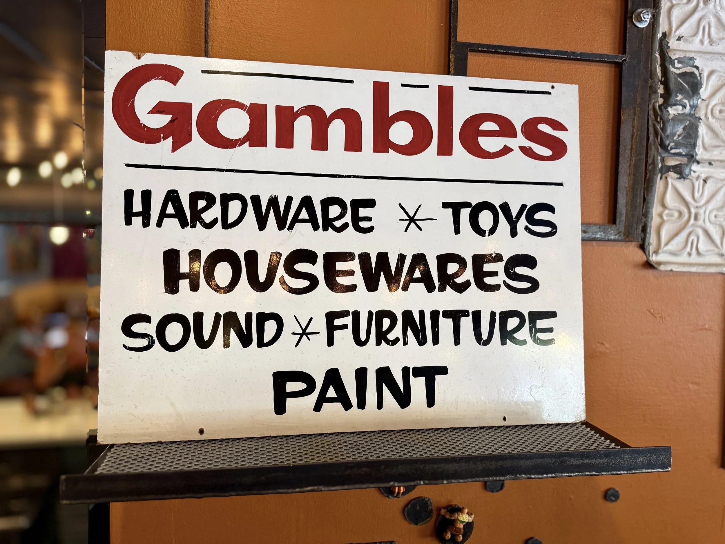 Sign with red and black text listing items for sale, including Gumballs, Hardware, Toys, Housewares, Sound, Furniture, and Paint, mounted on a wall with brown tiles and decorative molding.