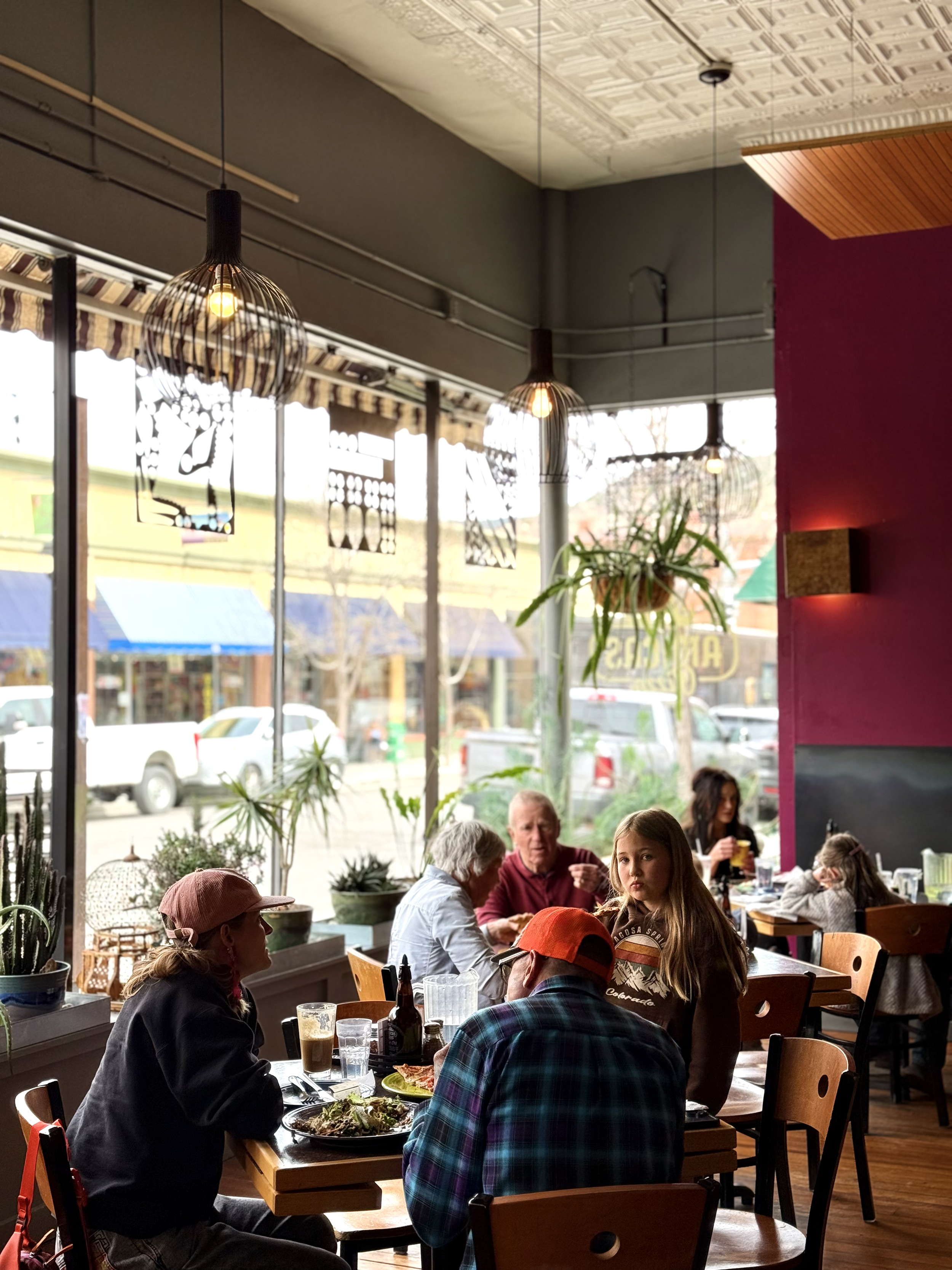 Interior of a busy restaurant with wooden tables and chairs, large window with a striped awning outside, hanging pendant lights, plants near the window, and several people dining and talking.