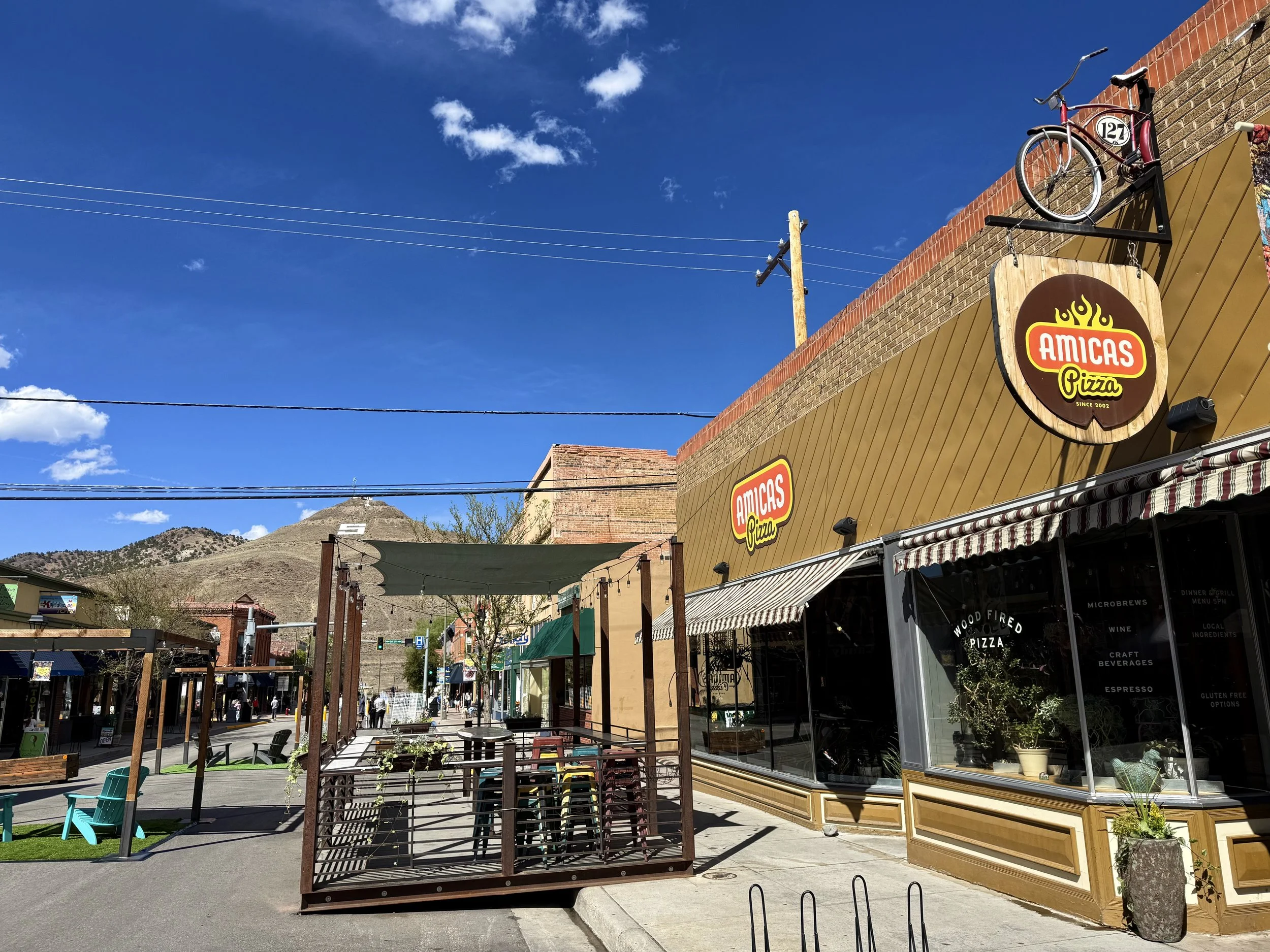 Street scene with a pizza restaurant named 'Amicas Pizza', outdoor seating area, and a bicycle mounted on the building's wall. Mountain in the background, blue sky with some clouds.