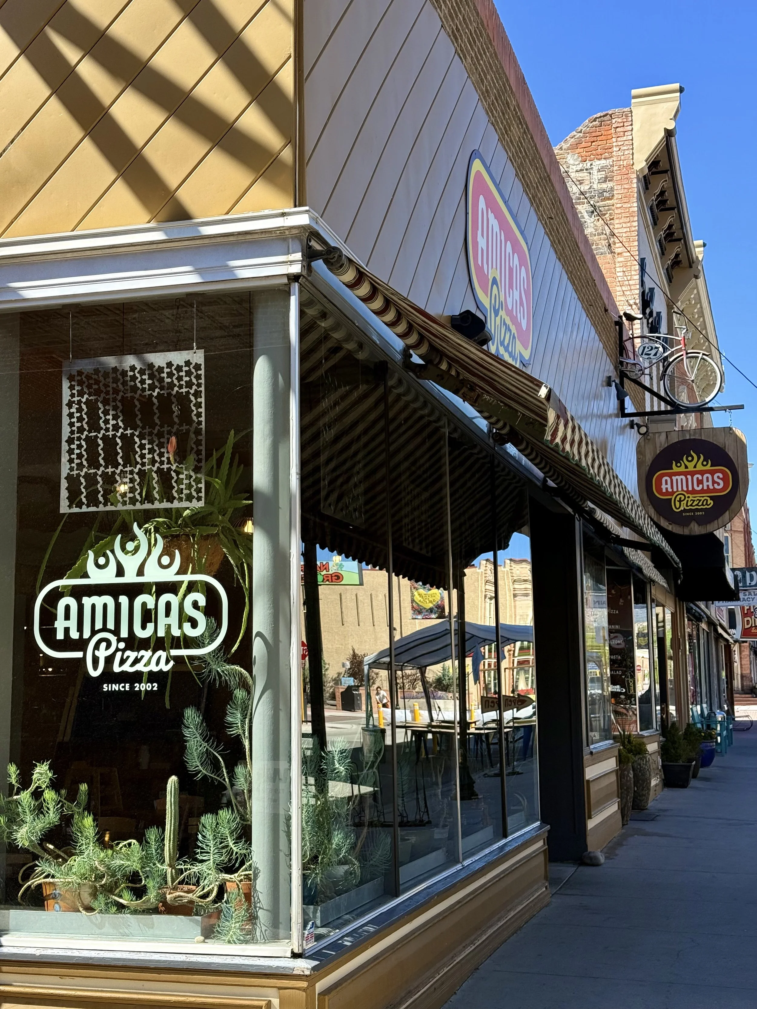 Outside view of a restaurant named Amicas Pizza, with a glass window at the front and a sign above. The window has potted plants inside. The building has a brick and wood exterior, with clear blue sky above.