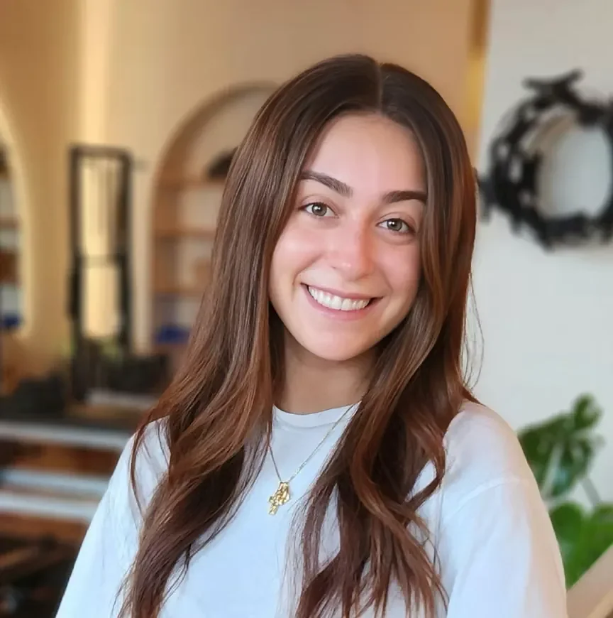 A young woman with long brown hair smiling indoors, wearing a white top and a gold necklace with a pendant.