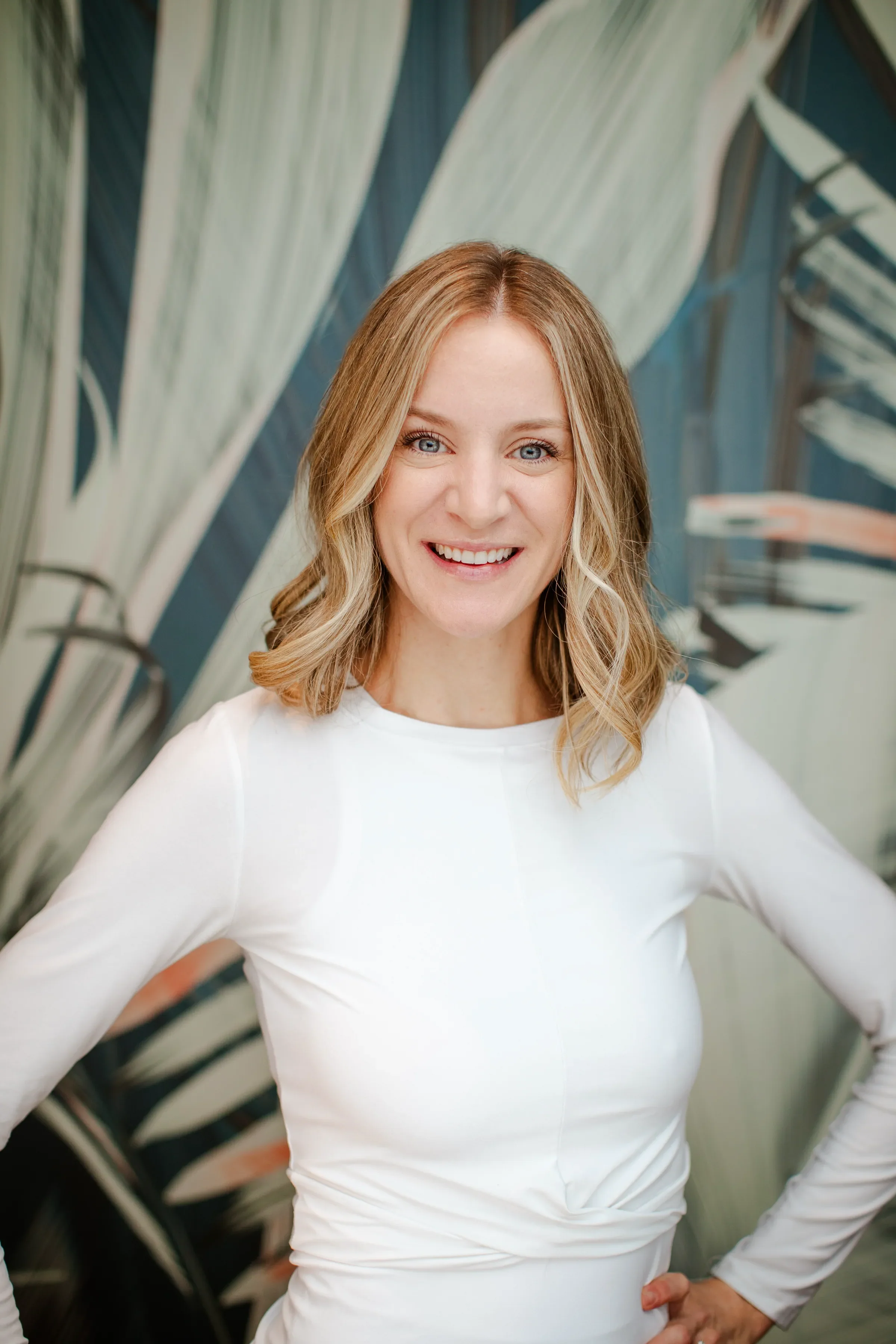 A woman with blonde hair wearing a white long-sleeve shirt, standing with her hands on her hips, smiling, with large leafy plants in the background.