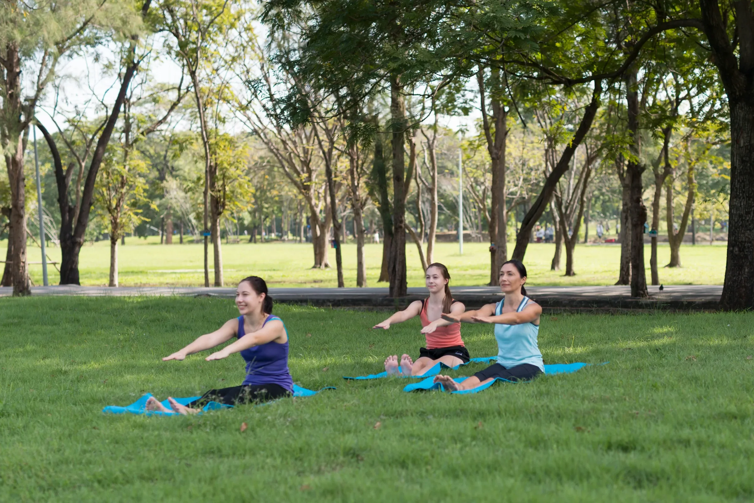 Three women practicing yoga outdoors on a grassy area in a park with tall trees in the background, sitting on blue yoga mats with their legs extended and arms reaching forward.