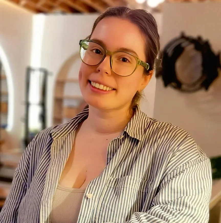A smiling woman with brown hair, wearing glasses, a beige tank top, and a striped button-down shirt, in a cozy indoor setting.