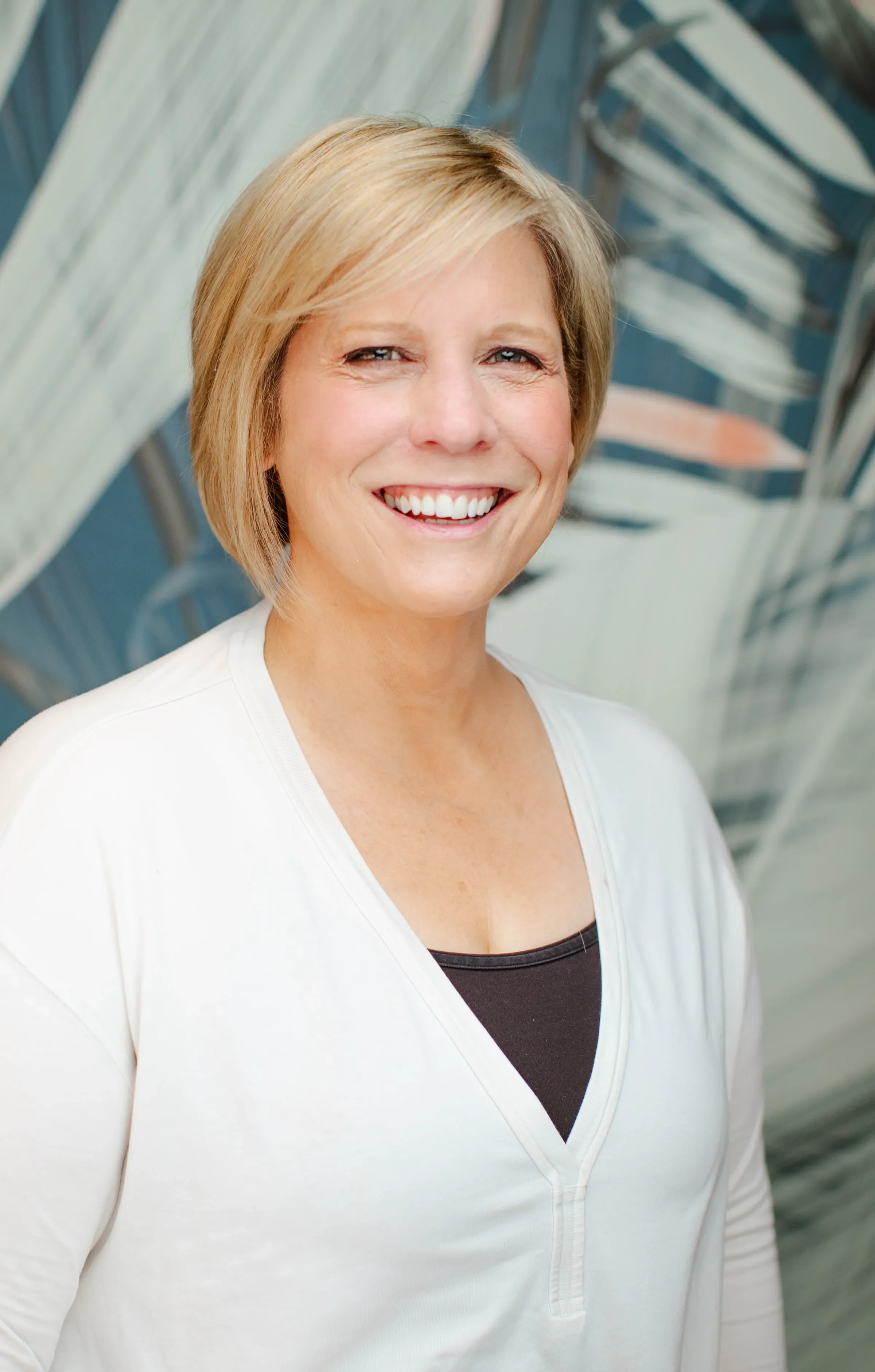 A smiling blonde woman with short hair, wearing a white top with a black shirt underneath, standing outdoors with a background of a glass building and metal structure.