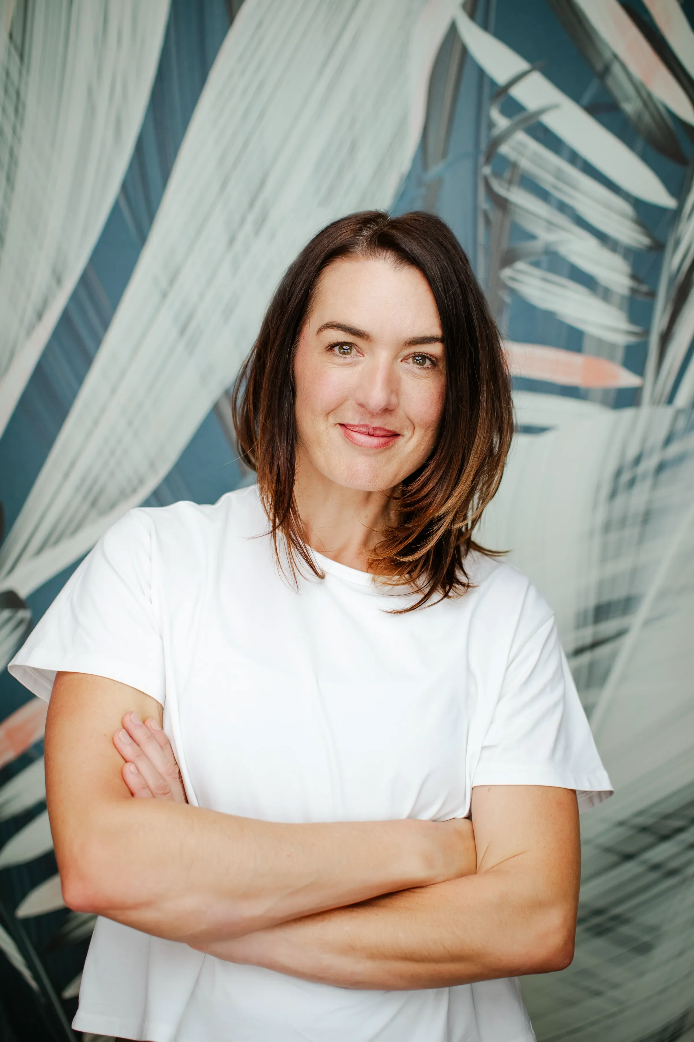 A woman with shoulder-length brown hair, wearing a white T-shirt, standing indoors in front of a wall decorated with abstract art of large white and blue leaves. She has a confident expression with arms crossed.