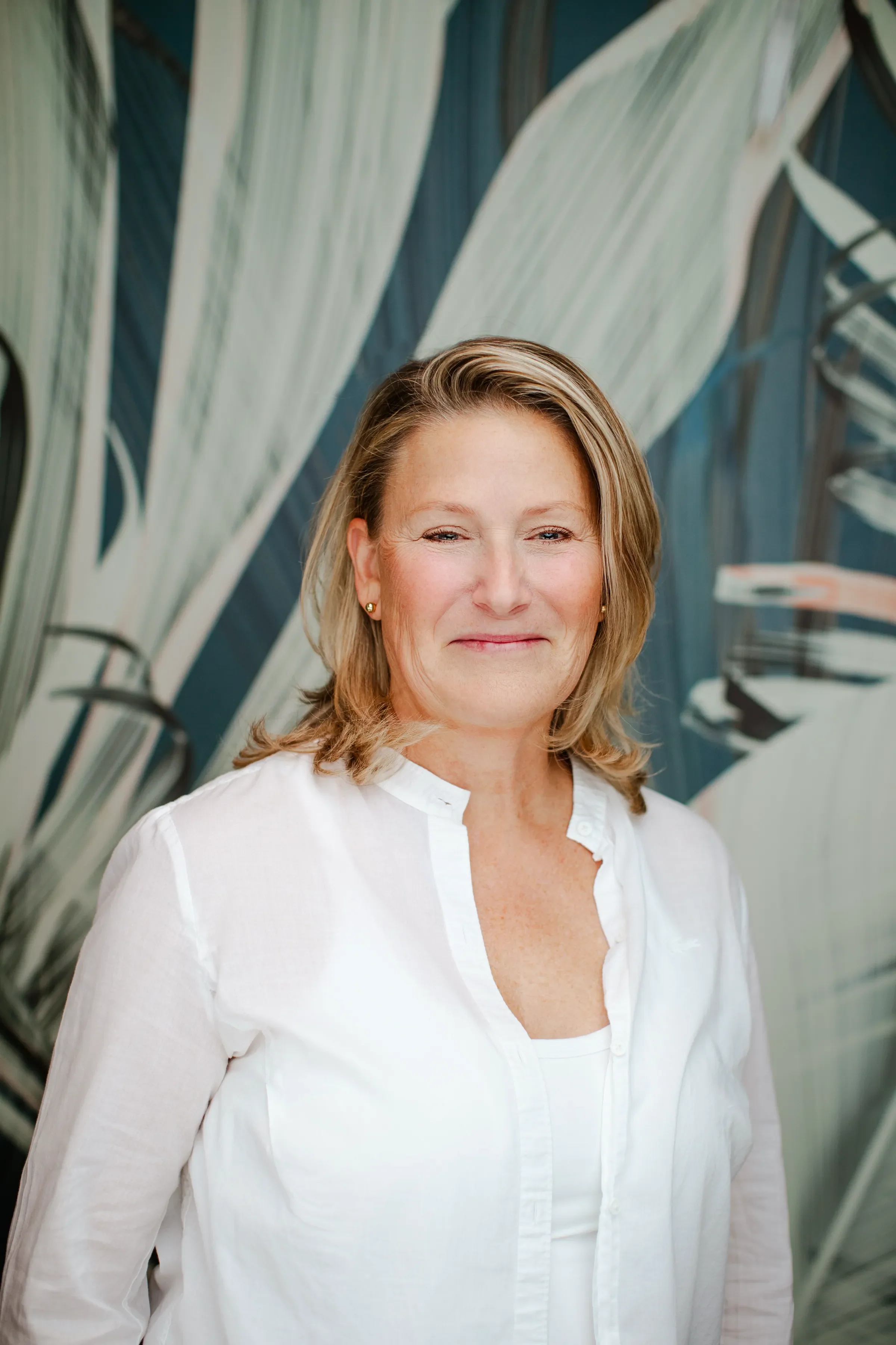 A middle-aged woman with blonde hair and earrings, wearing a white button-down shirt, standing in front of a background with large tropical leaves.