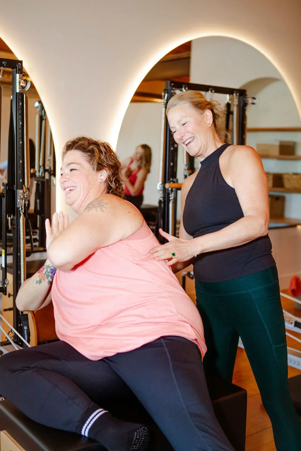 Two women exercising and laughing in a fitness studio, one sitting on a workout bench and the other standing, both smiling.