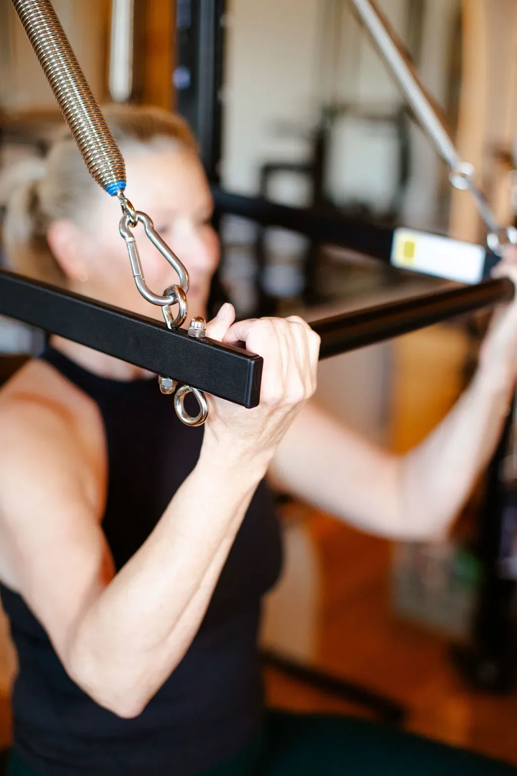 A woman is doing a shoulder exercise on a cervical pulley machine in a gym, with her face blurred out.