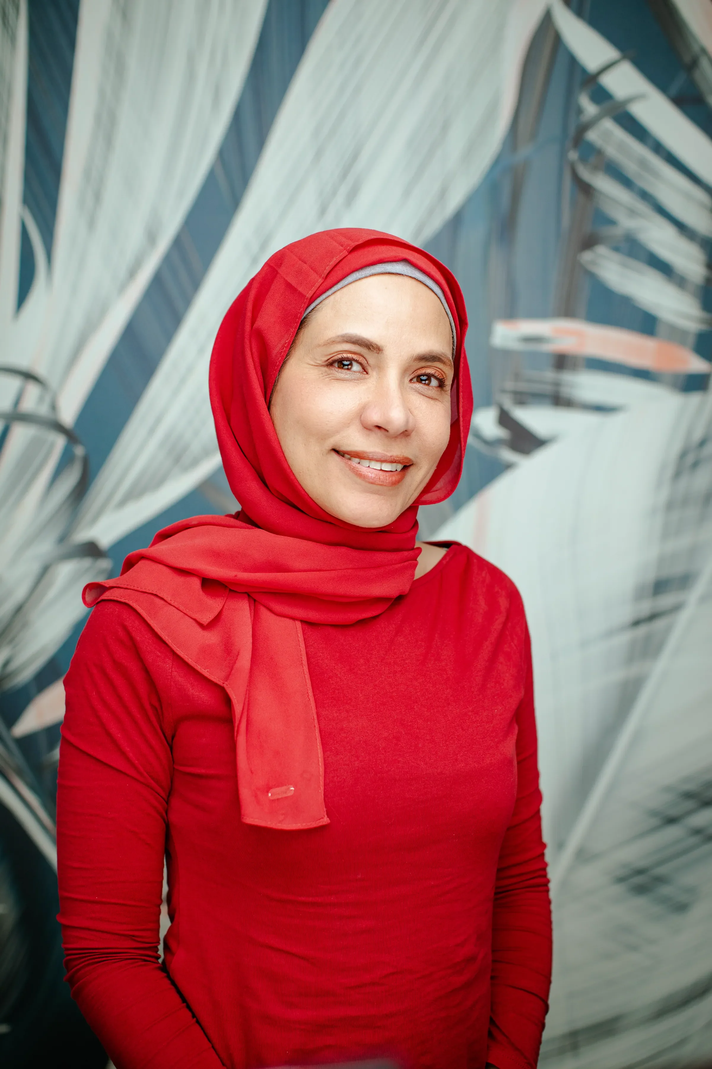 A woman wearing a red hijab and matching red top standing in front of a wall of what appears to be metal or glass structures in an artistic or modern architectural setting.