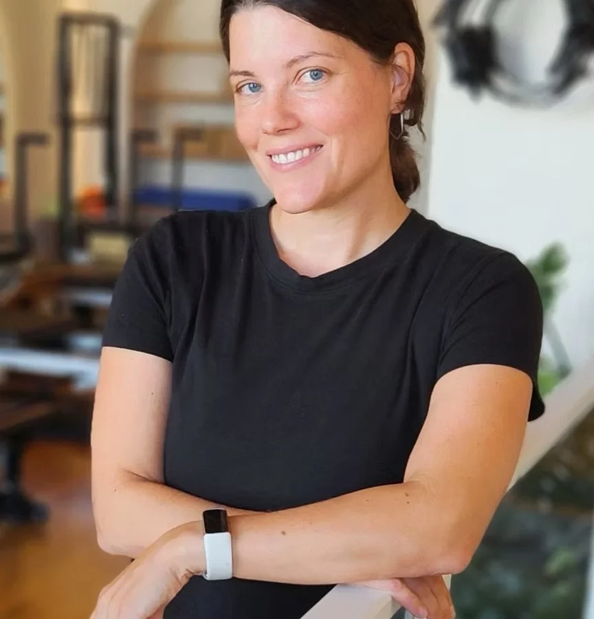 A woman with short brown hair and blue eyes wearing a black t-shirt, smiling with arms crossed in a cozy home setting.