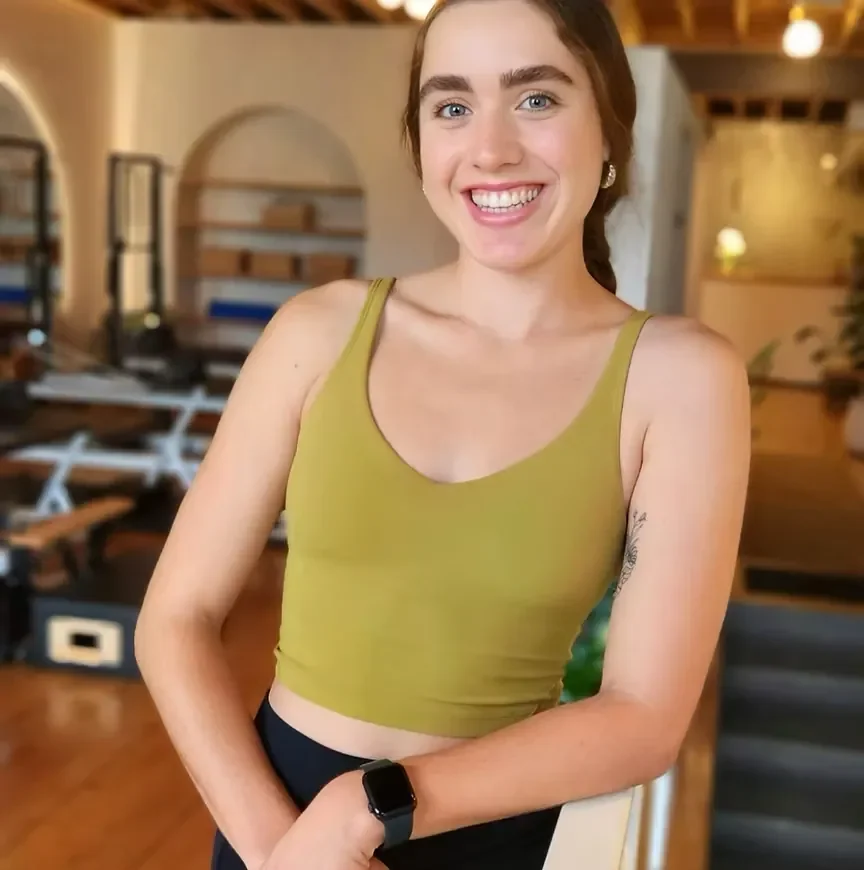 A young woman with brown hair tied back, smiling, wearing a green sleeveless crop top, and an Apple Watch on her left wrist, standing in a cozy, well-lit indoor space with wooden furniture and plants.