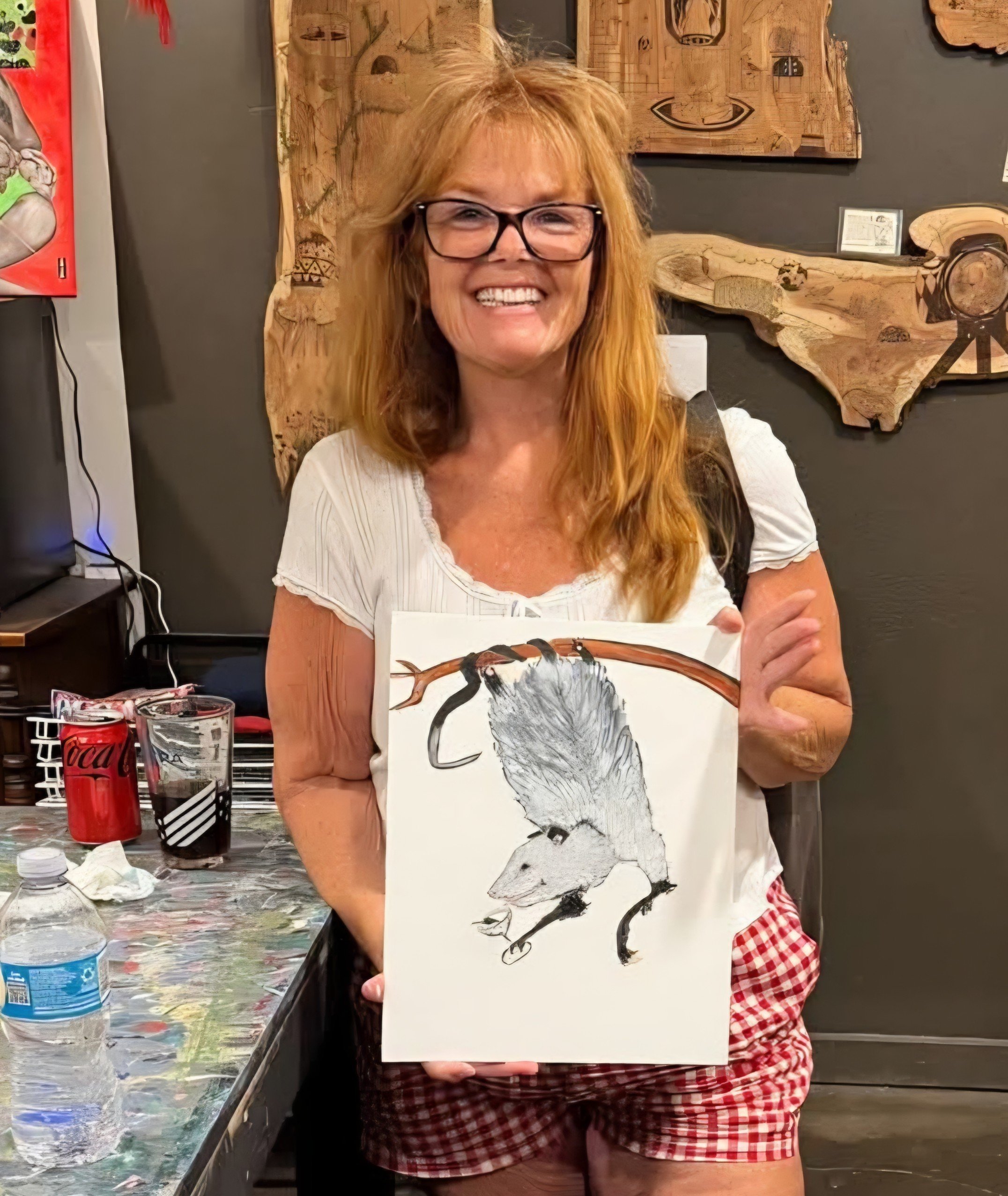 Woman with glasses smiling and holding a painting of a bird on canvas, standing in front of wooden art pieces on wall in an art studio.