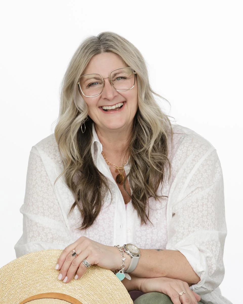A smiling middle-aged woman with long wavy blonde hair, glasses, wearing a white blouse with lace, gold jewelry, and a wristwatch, holding a straw hat against a white background.