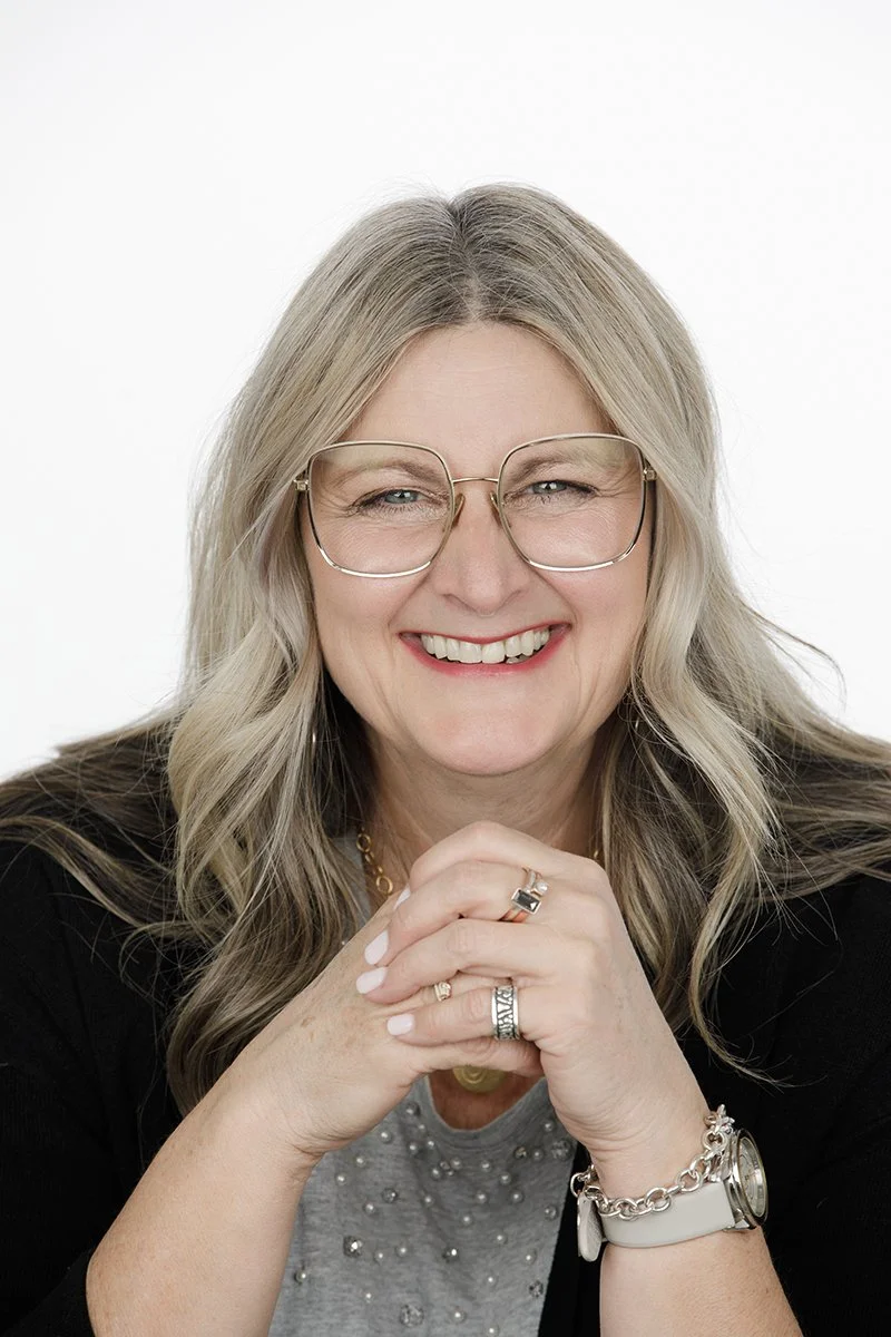 Portrait of a smiling woman with long blonde hair, wearing glasses, rings, a bracelet, and a watch, against a white background.