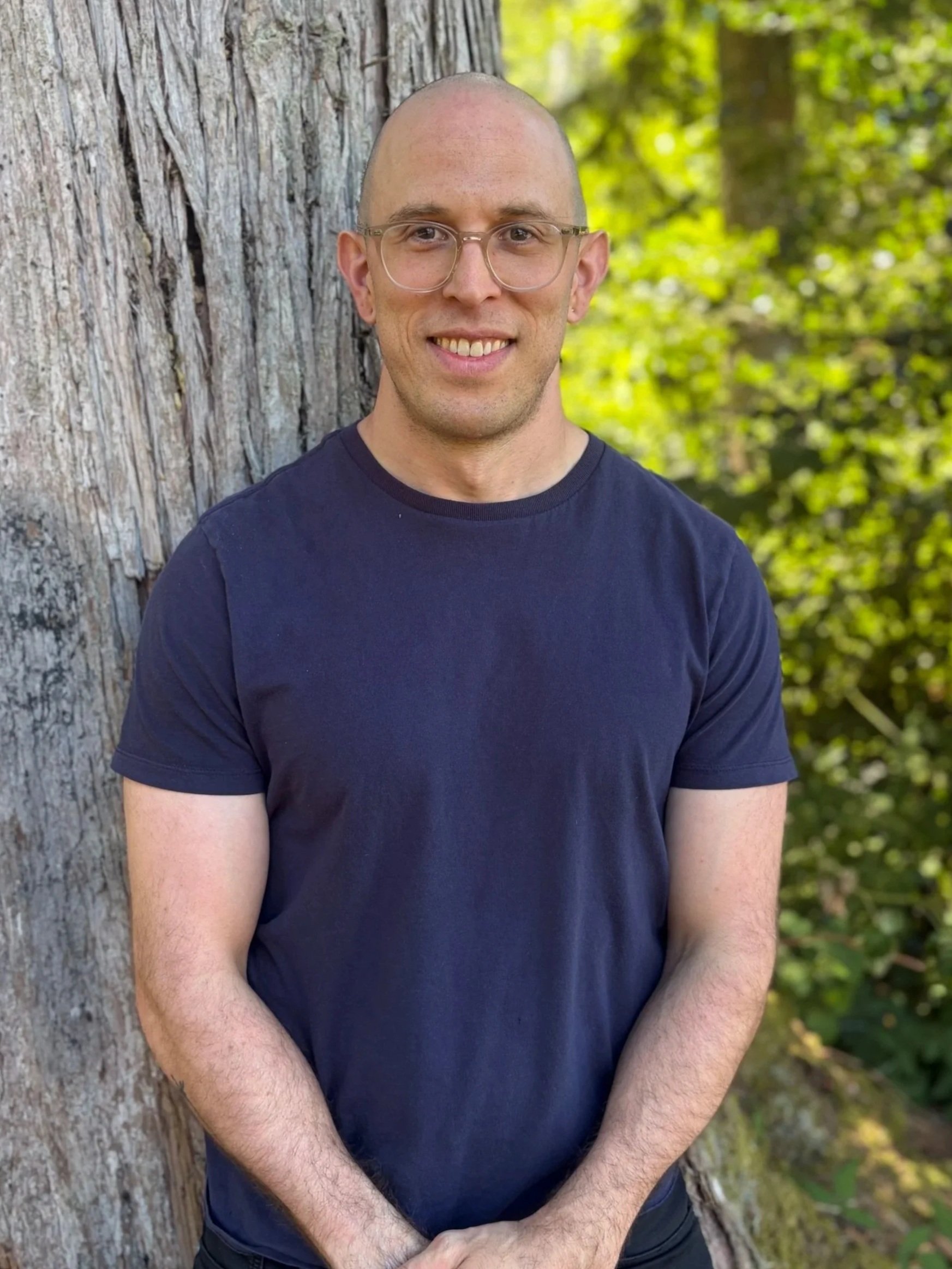 A man with glasses and a shaved head smiling, standing outdoors next to a large tree with green leaves in the background.