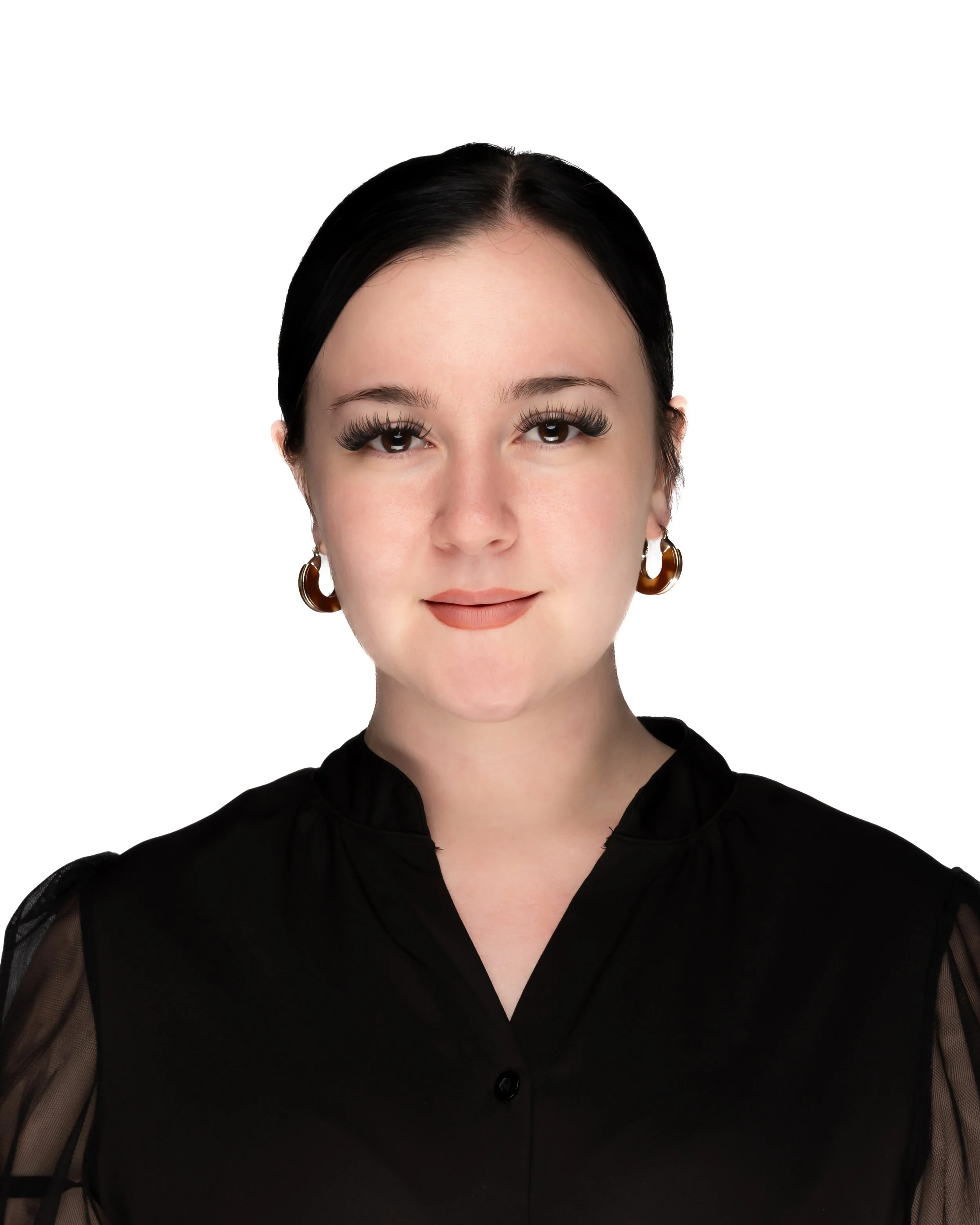 Close-up of a woman with dark hair styled back, wearing large hoop earrings, a black sheer blouse with puffed sleeves, and natural makeup with defined eyelashes, against a white background.