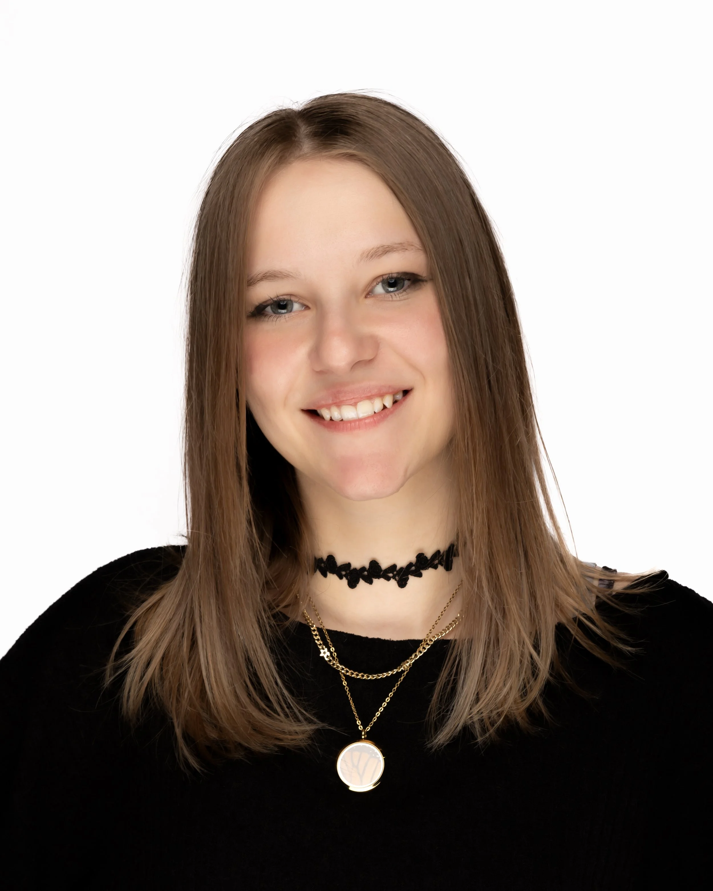 Close-up of a smiling young woman with long light brown hair, wearing a black top, layered necklaces including a circular pendant, a black lace choker, and layered chains, against a white background.