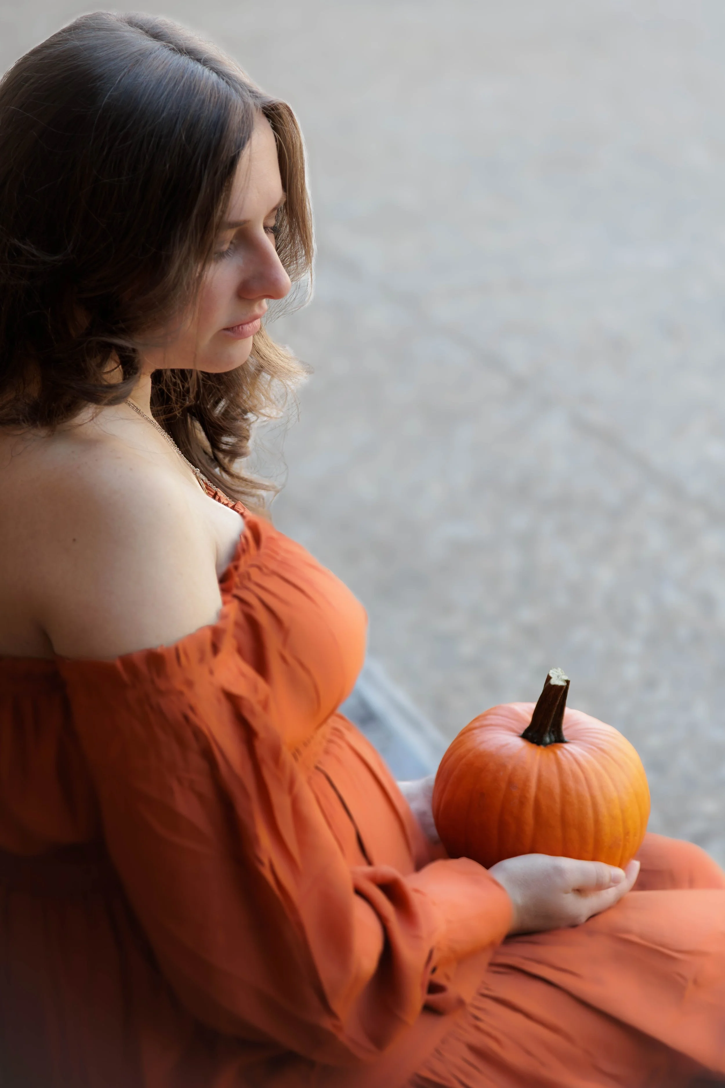 A woman with brown hair in an orange off-shoulder dress holding a small pumpkin with a stem, sitting near a body of water.