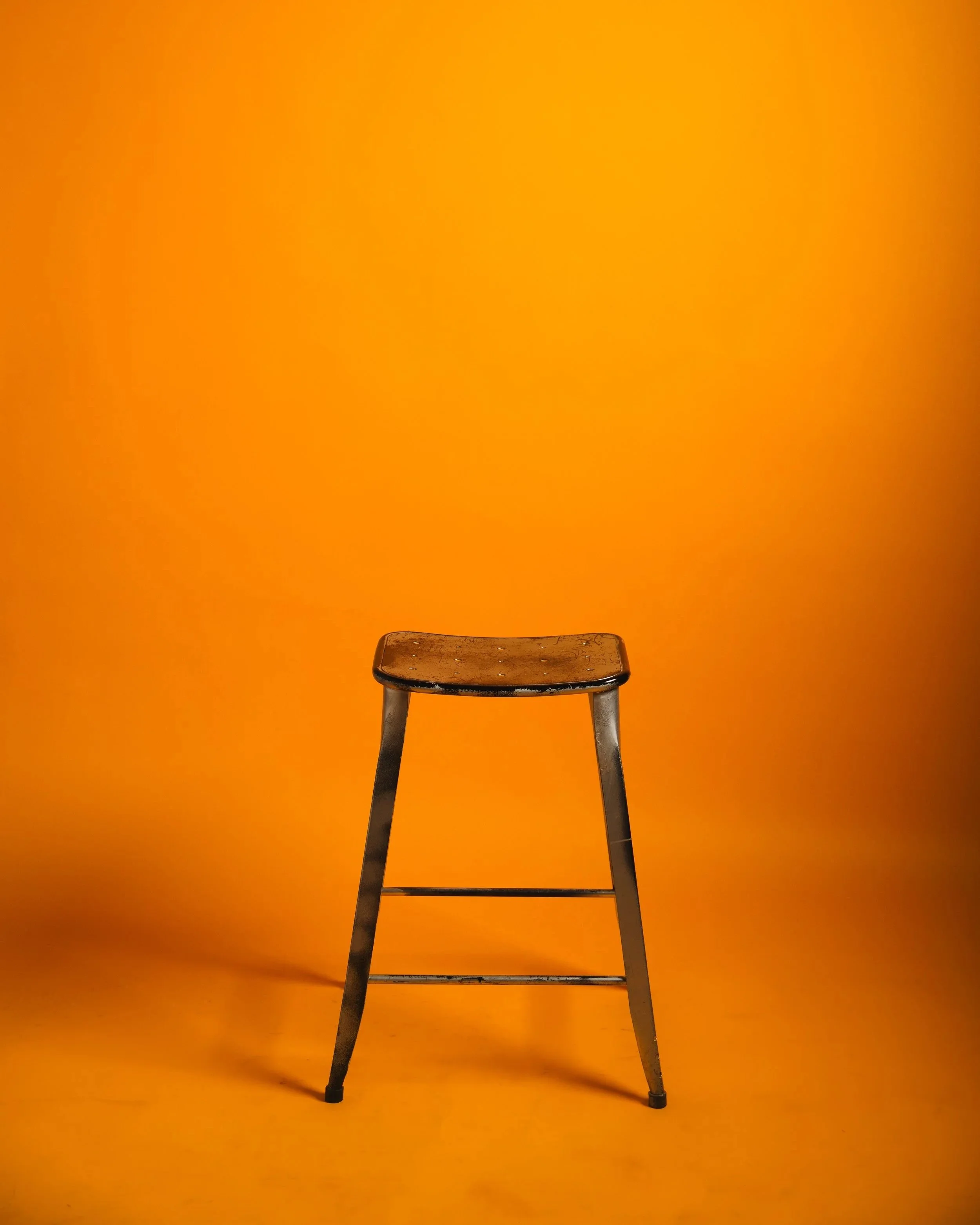 An old wooden stool with gray metal legs against an orange background.
