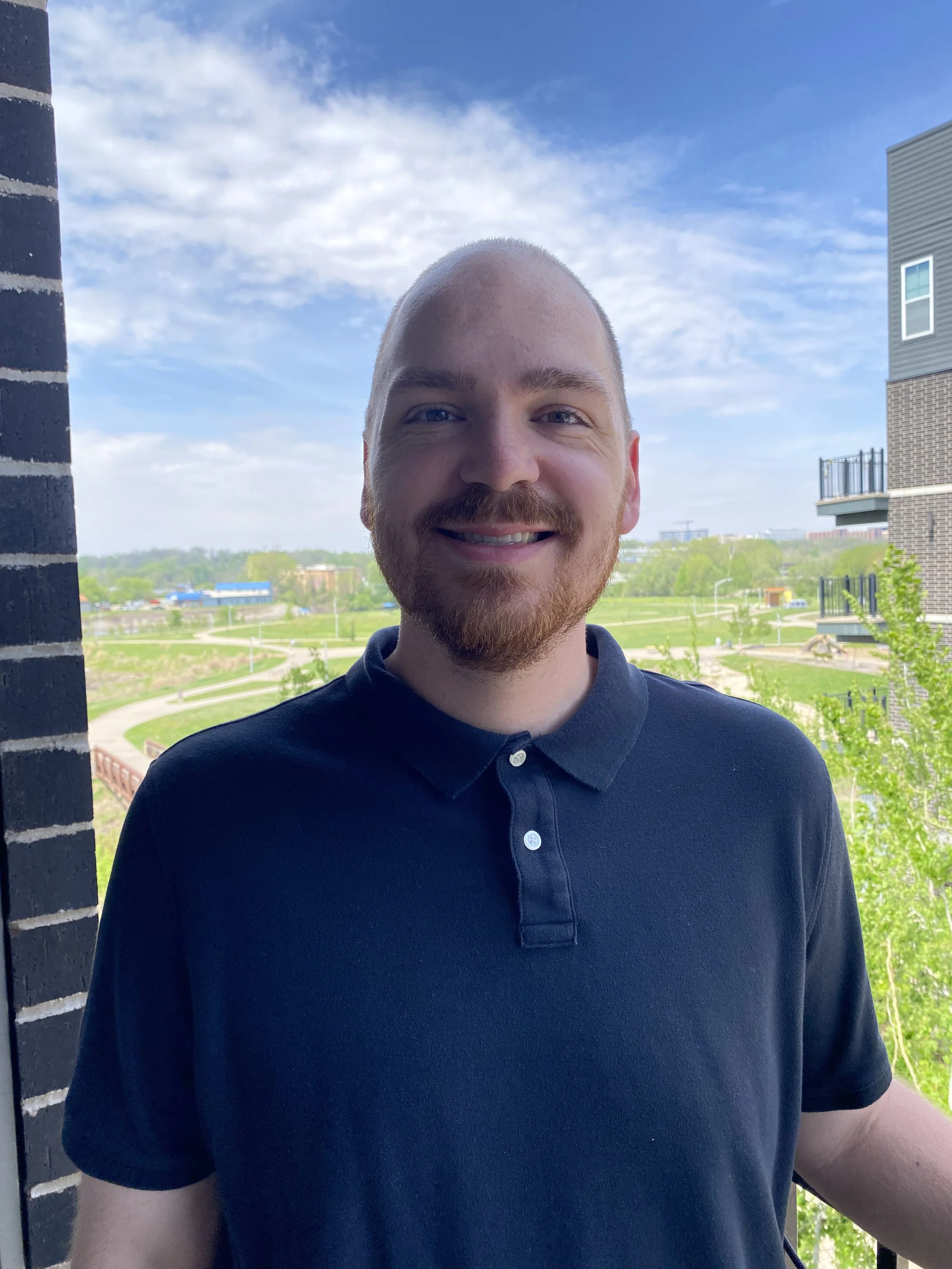 A young man with short hair and a beard smiling, standing outside near a brick wall with a view of a park, buildings, and a partly cloudy sky in the background.