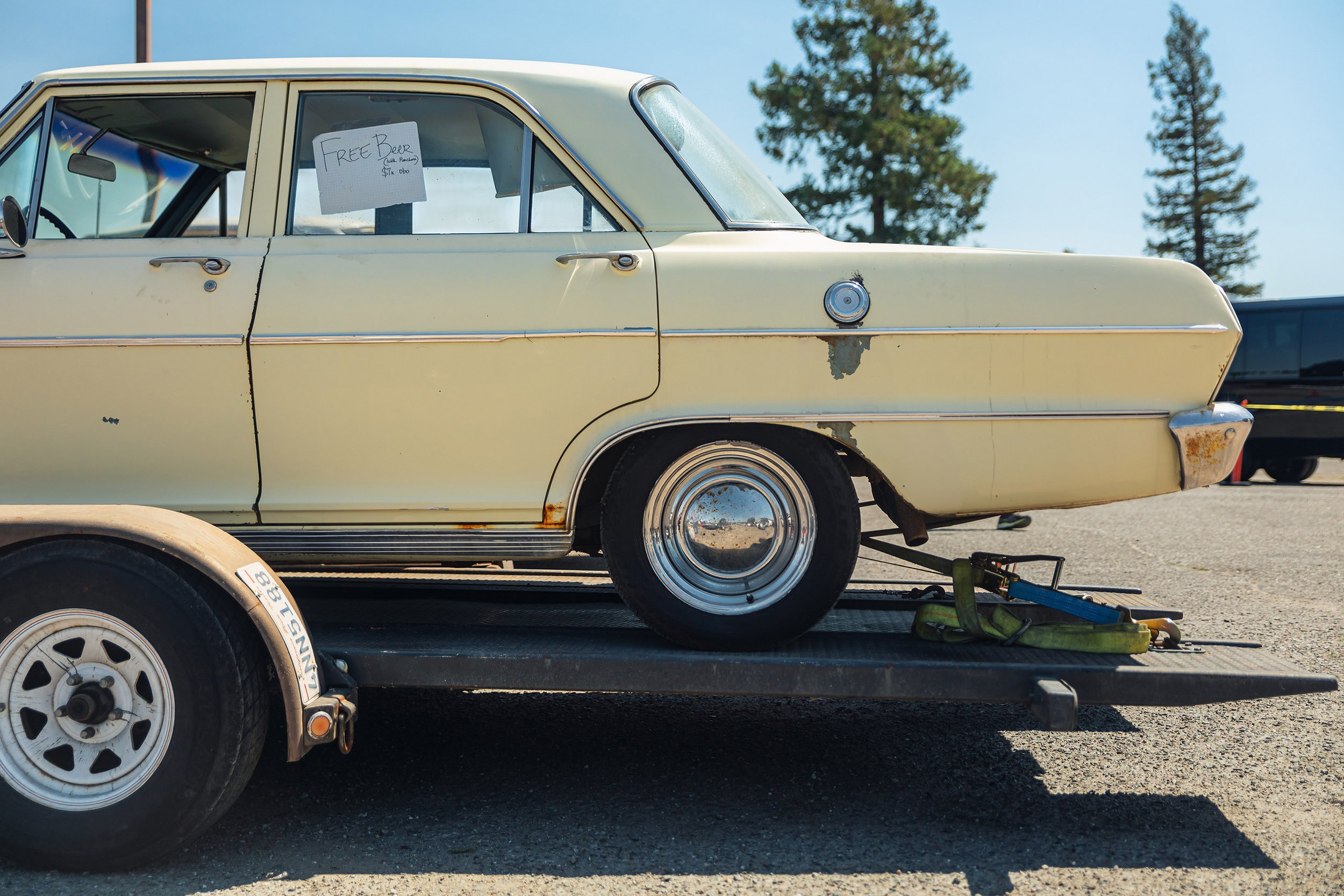Vintage yellow car on a trailer with a sign in the window offering free beer, parked outdoors with trees in the background.