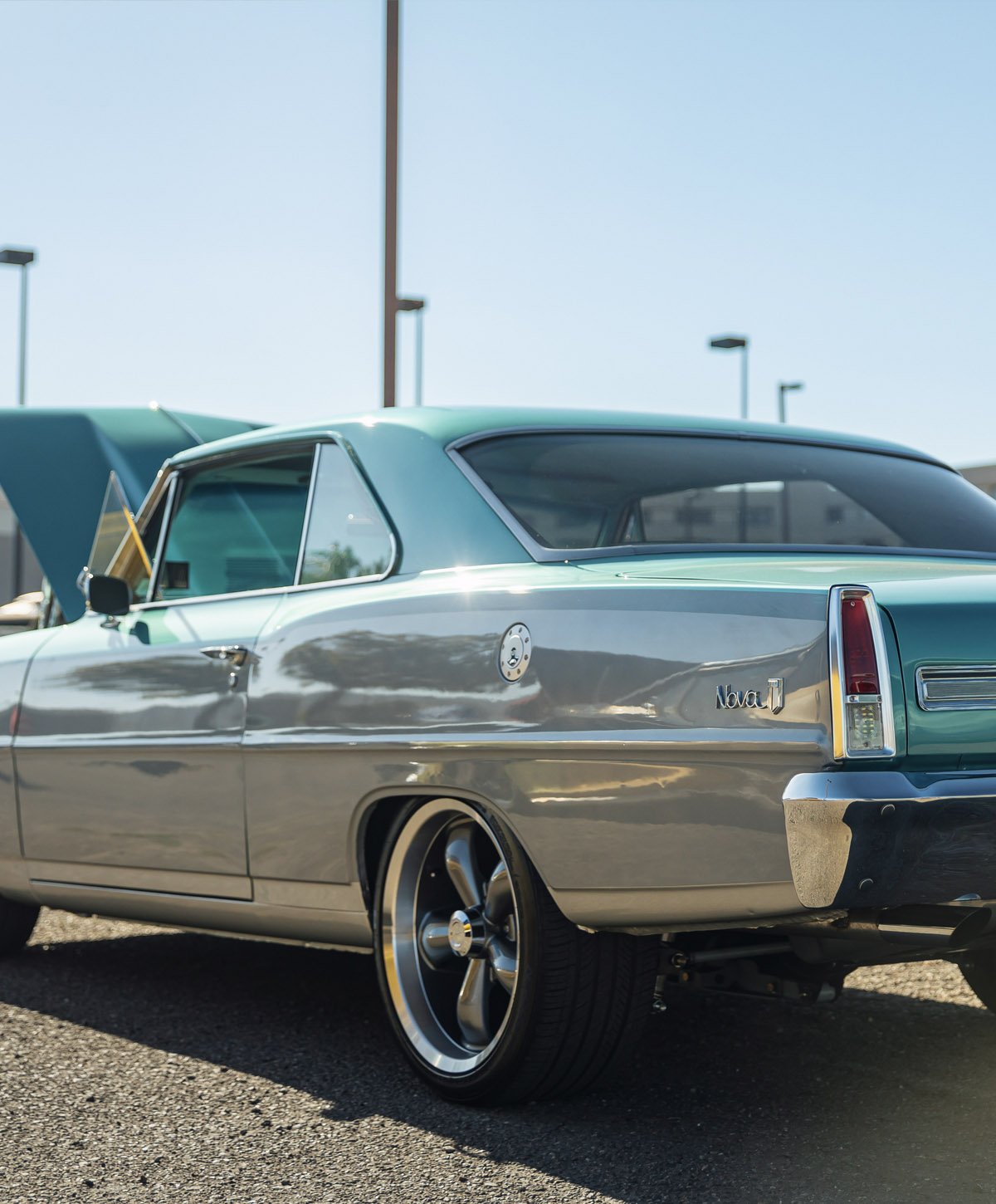 A vintage two-tone blue and silver Chevrolet Nova parked in a lot with a clear, sunny sky and lamp posts in the background.