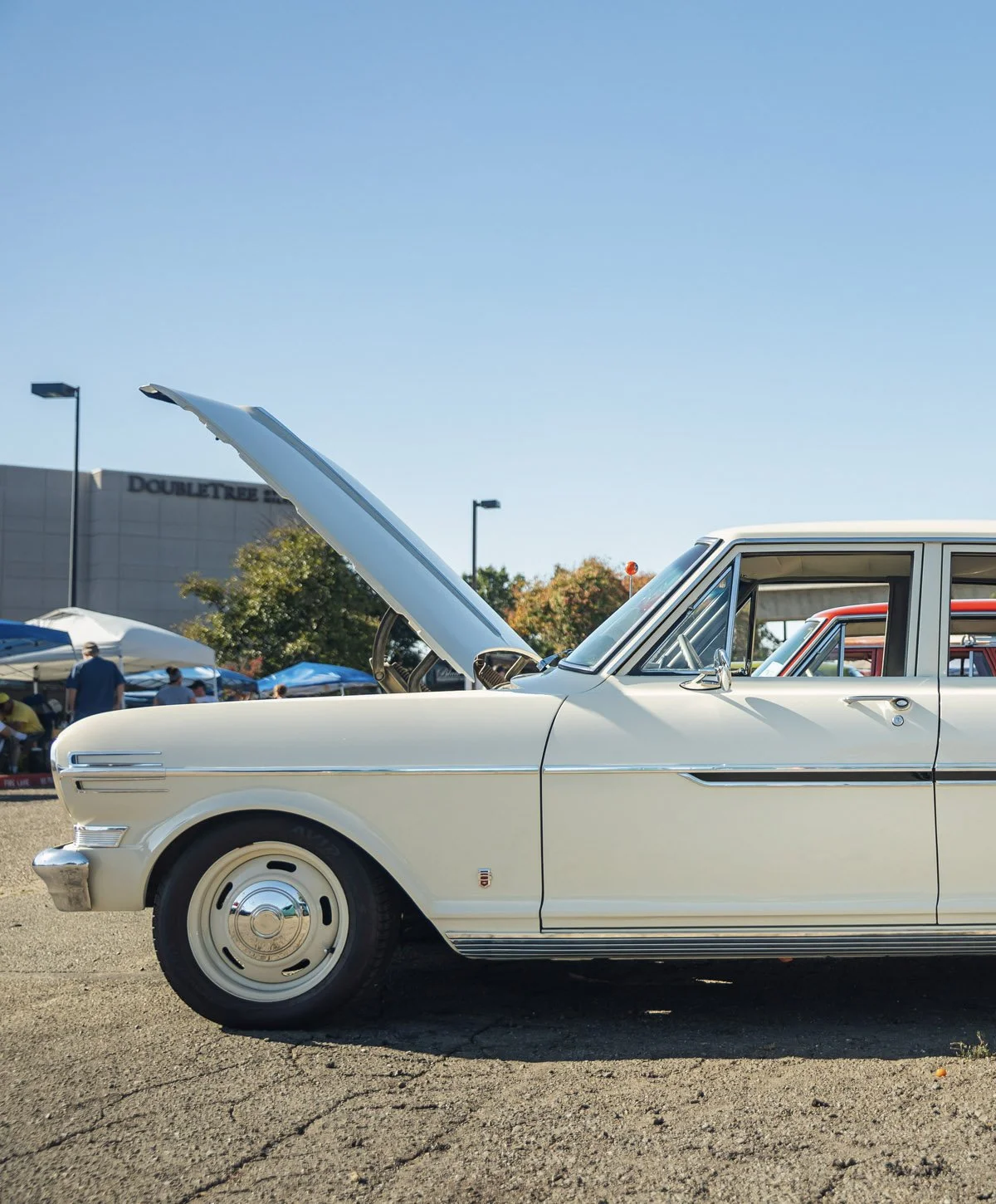 A vintage white car with an open hood at a car show, with tents and people in the background and a DoubleTree hotel building visible.