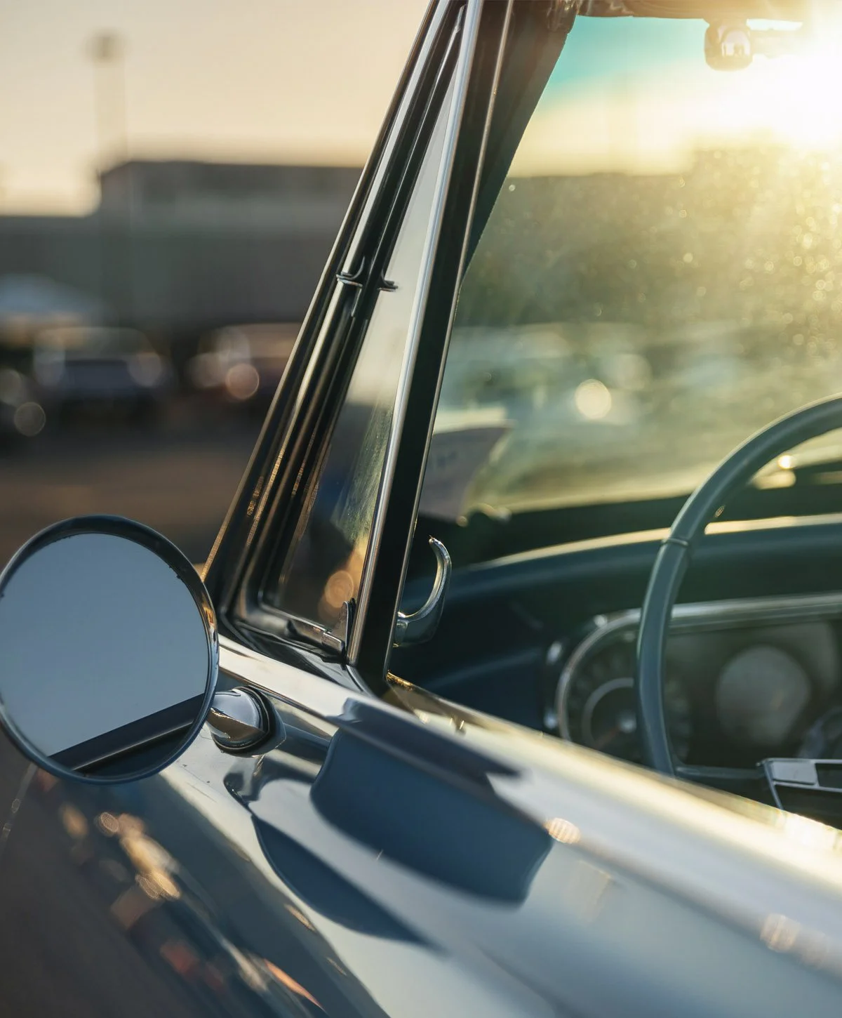 Close-up of the front part of a vintage car showing the side mirror, windshield, and steering wheel with sunlight in the background.