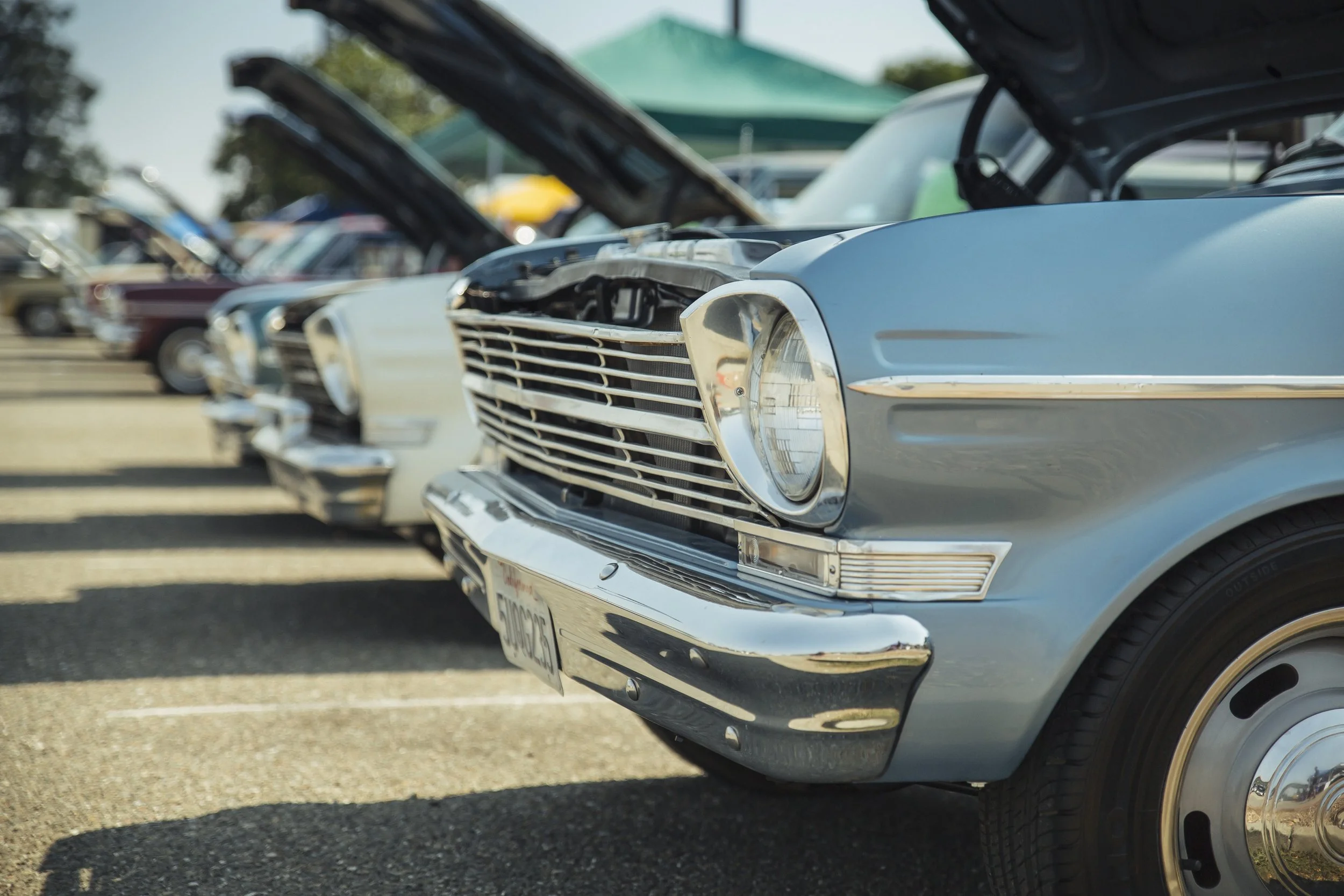 Row of classic cars at a car show, with open hoods, parked on a lot with trees in the background.