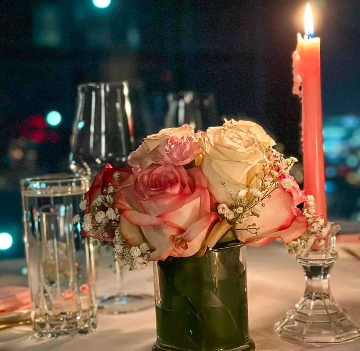 A table decorated with a floral centerpiece of pink and white roses, a lit pink candle, glasses of water, and a city skyline visible in the background at night.