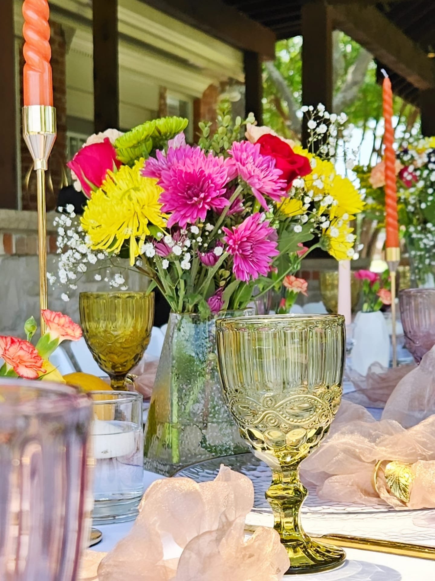 A table decorated with colorful flowers in various glass vases, surrounded by candles and light-colored fabric, outdoors with a house and trees in the background.