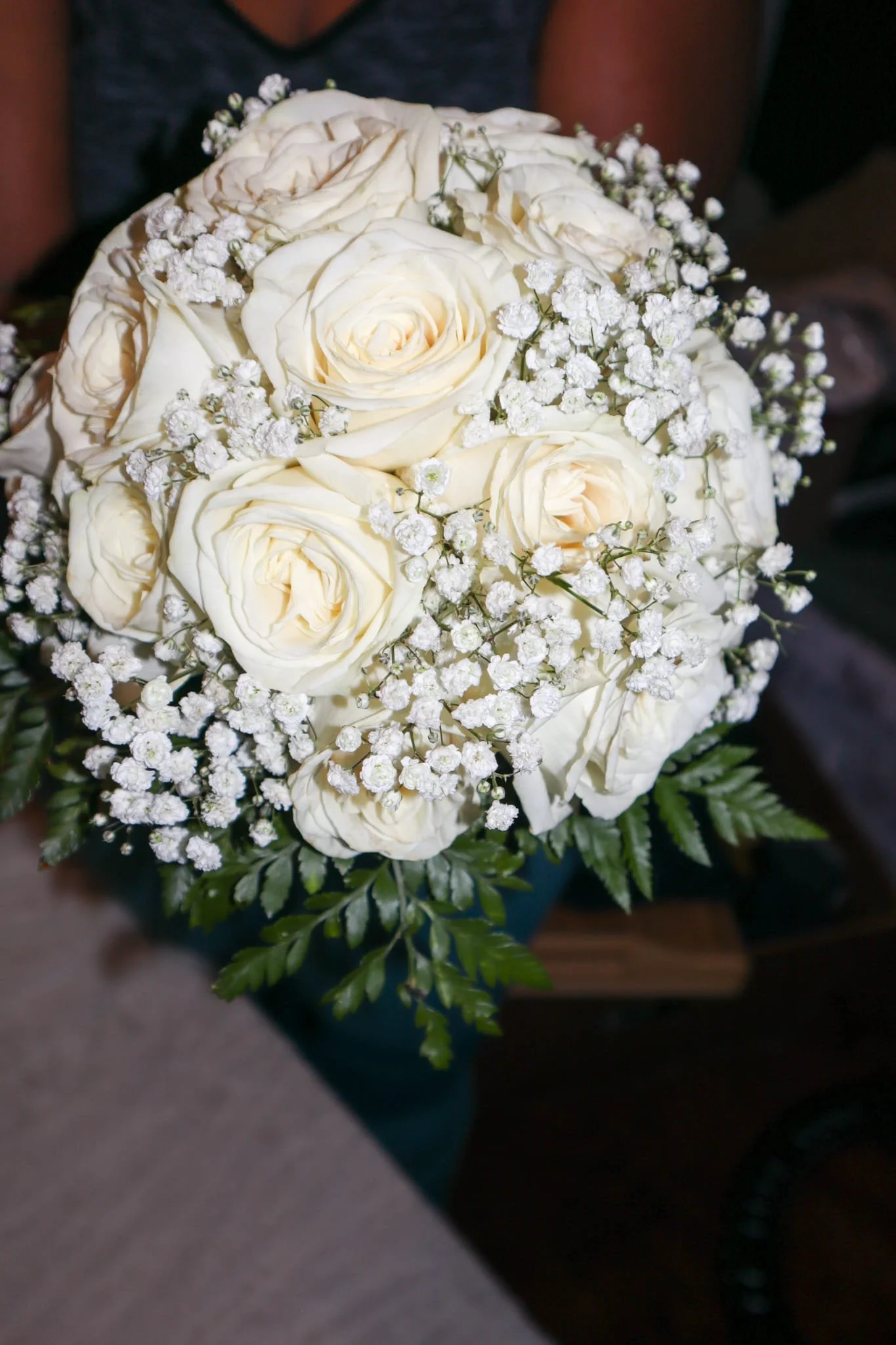 A bouquet of white roses with baby's breath flowers and green foliage, held by a person wearing a dark shirt.