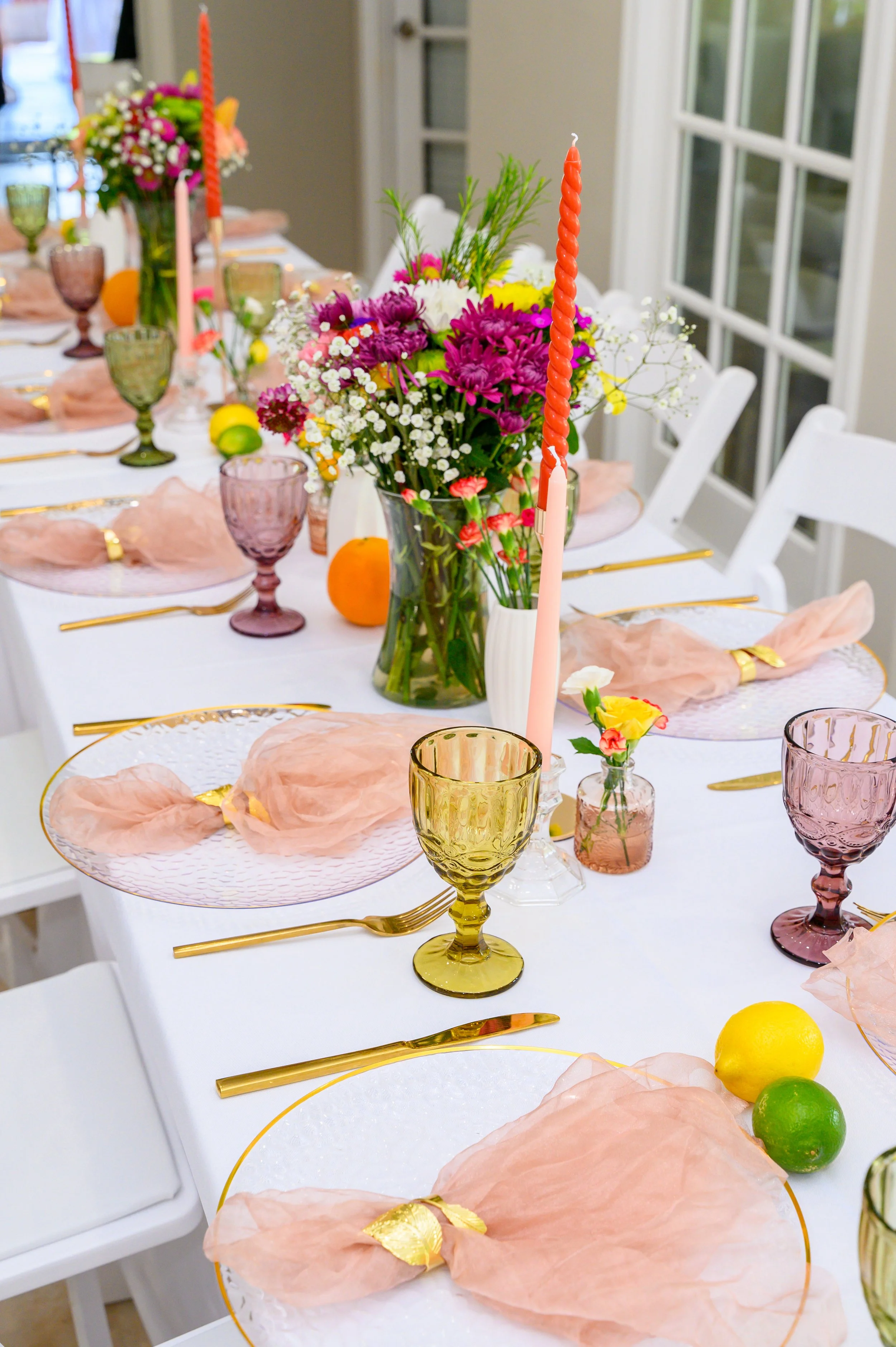 A decorated dining table with pastel pink napkins tied with gold rings, colorful flower arrangements in glass vases, tall textured candles, and colorful glasses. Whole lemons and limes are scattered on the table.
