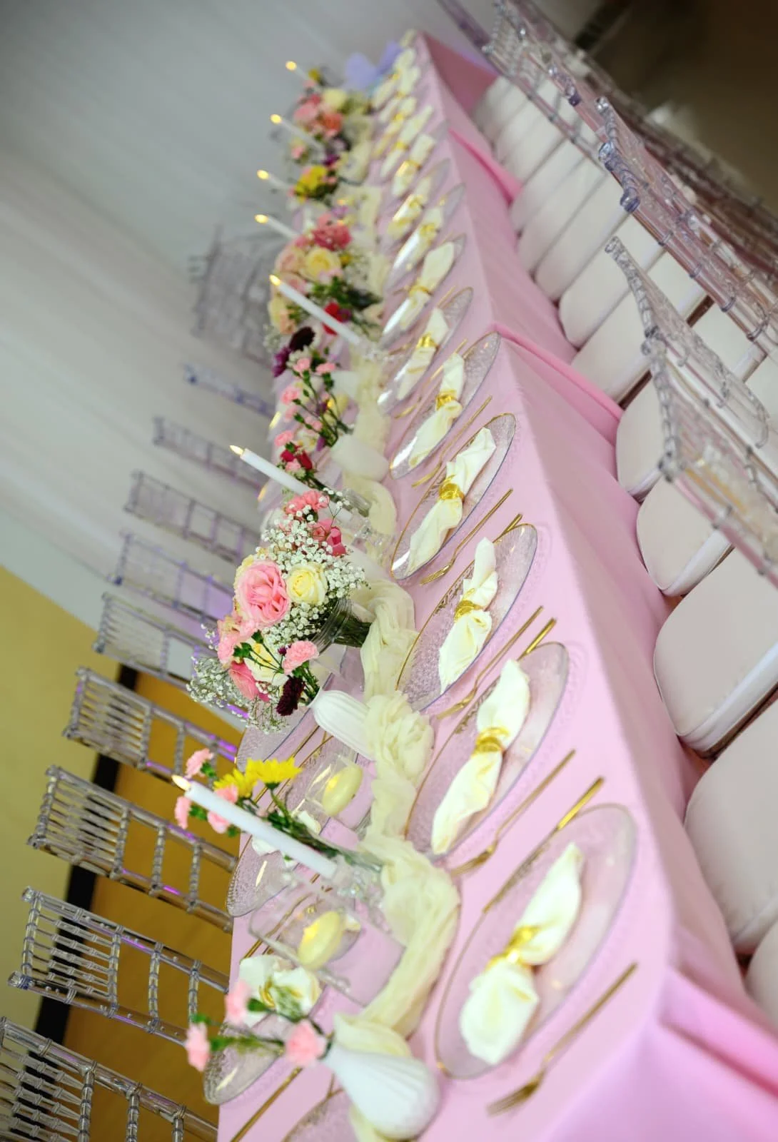 A long pink table with floral centerpieces in small jars, candles, and elegant place settings with gold cutlery and white napkins.