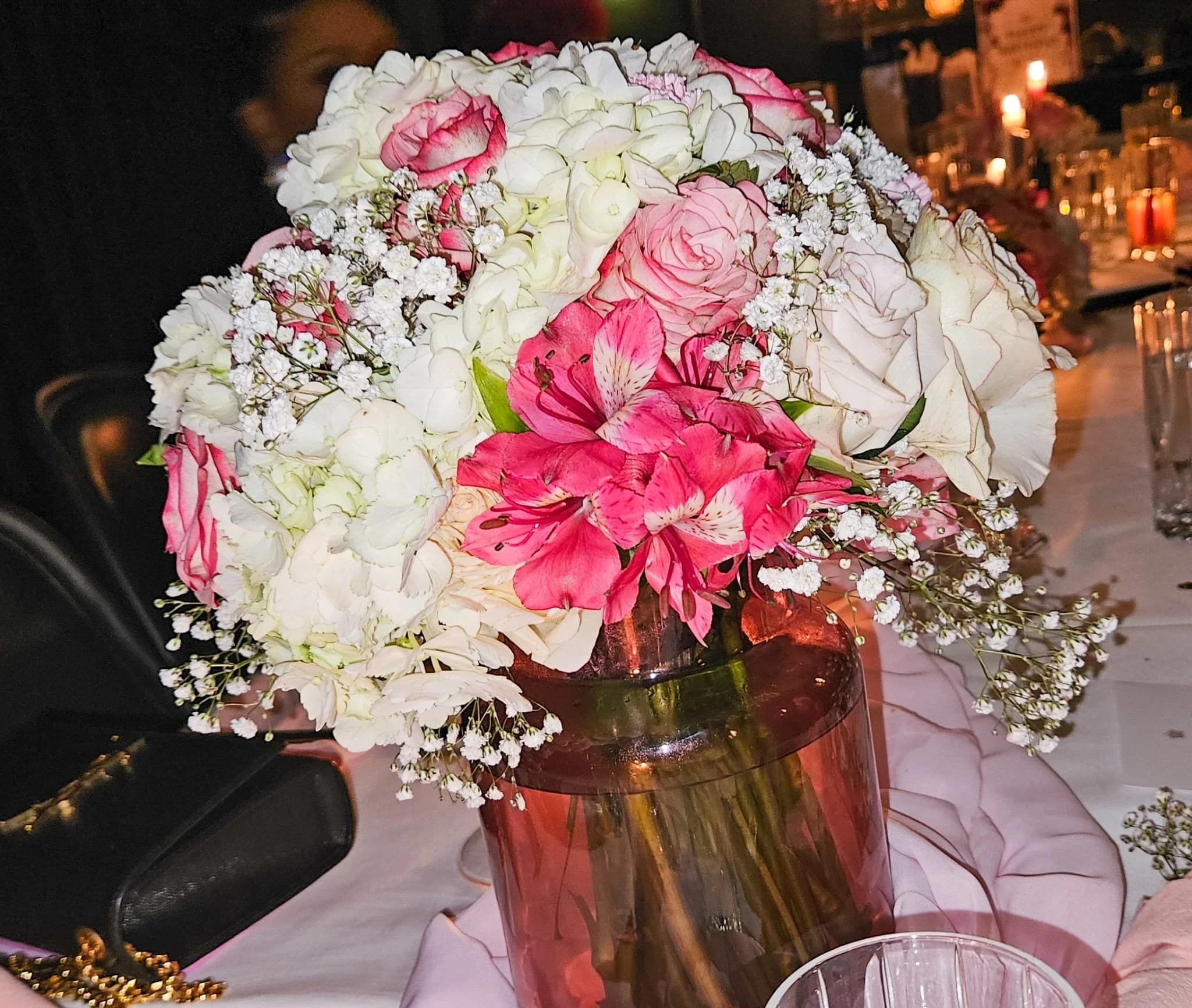 Close-up of a bouquet of pink and white flowers in a glass vase on a table, with blurred background lighting.