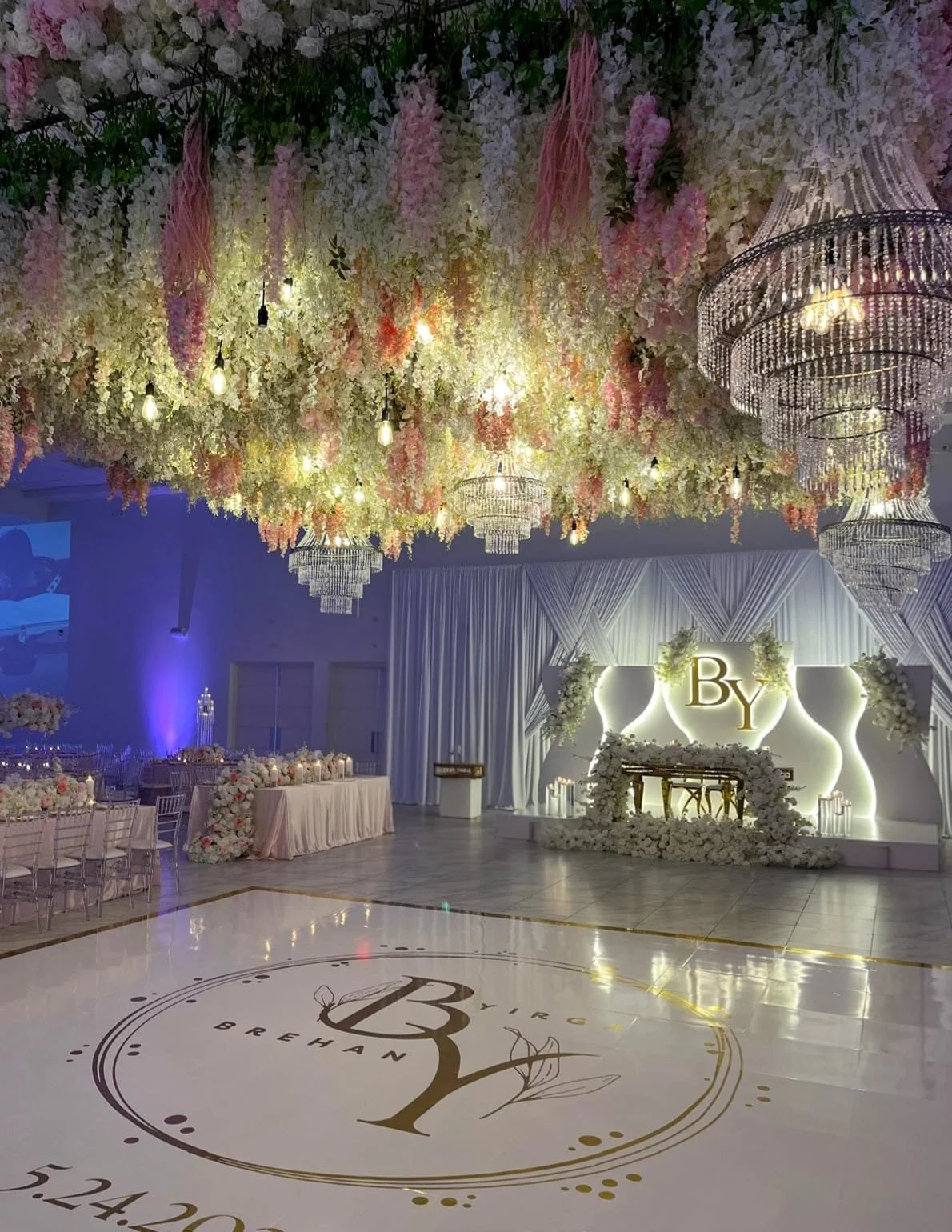 Wedding reception hall decorated with hanging flowers, pink and white floral arrangements, chandeliers, illuminated backdrop with initials 'B Y', and tables with flower centerpieces.