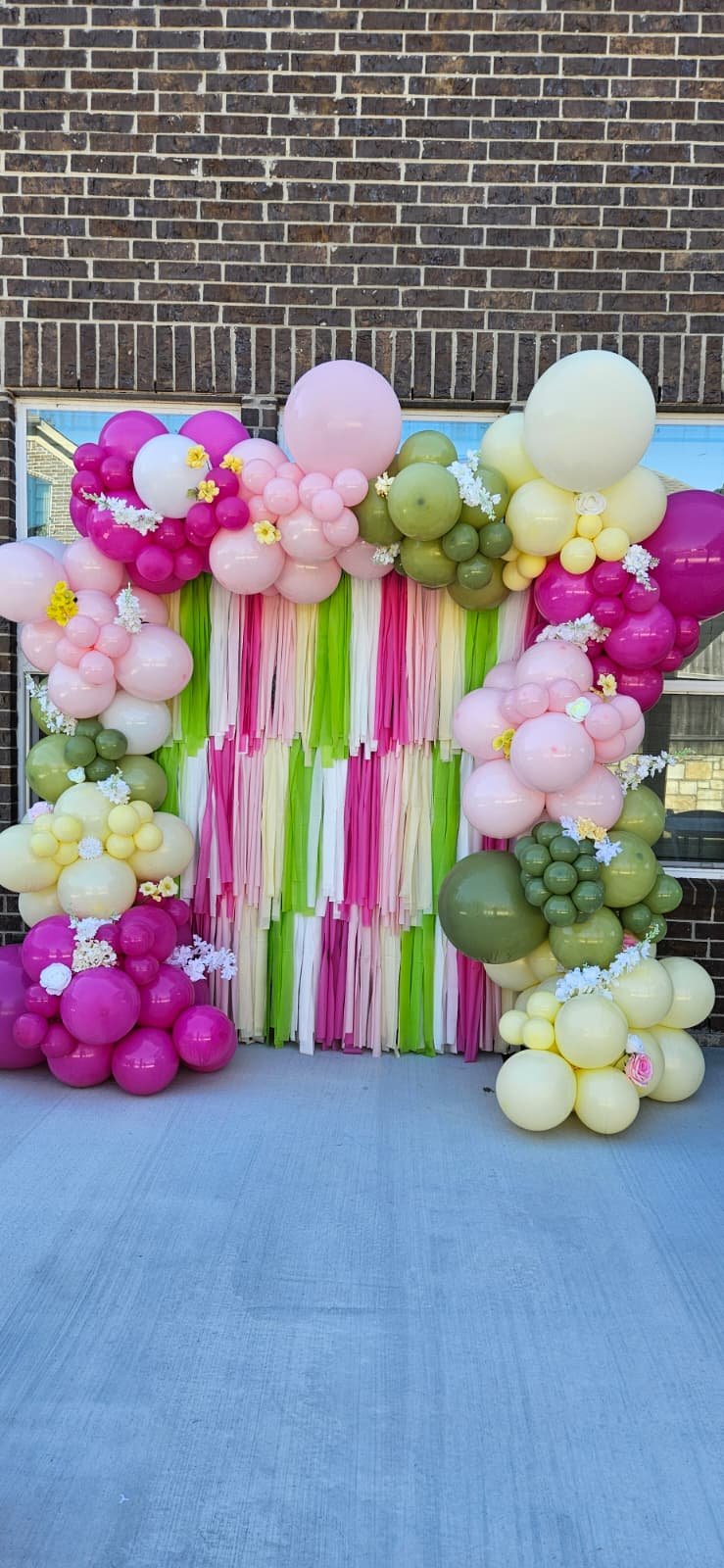 Decorative balloon arch with pink, green, yellow, and white balloons and streamers, set against a brick wall.