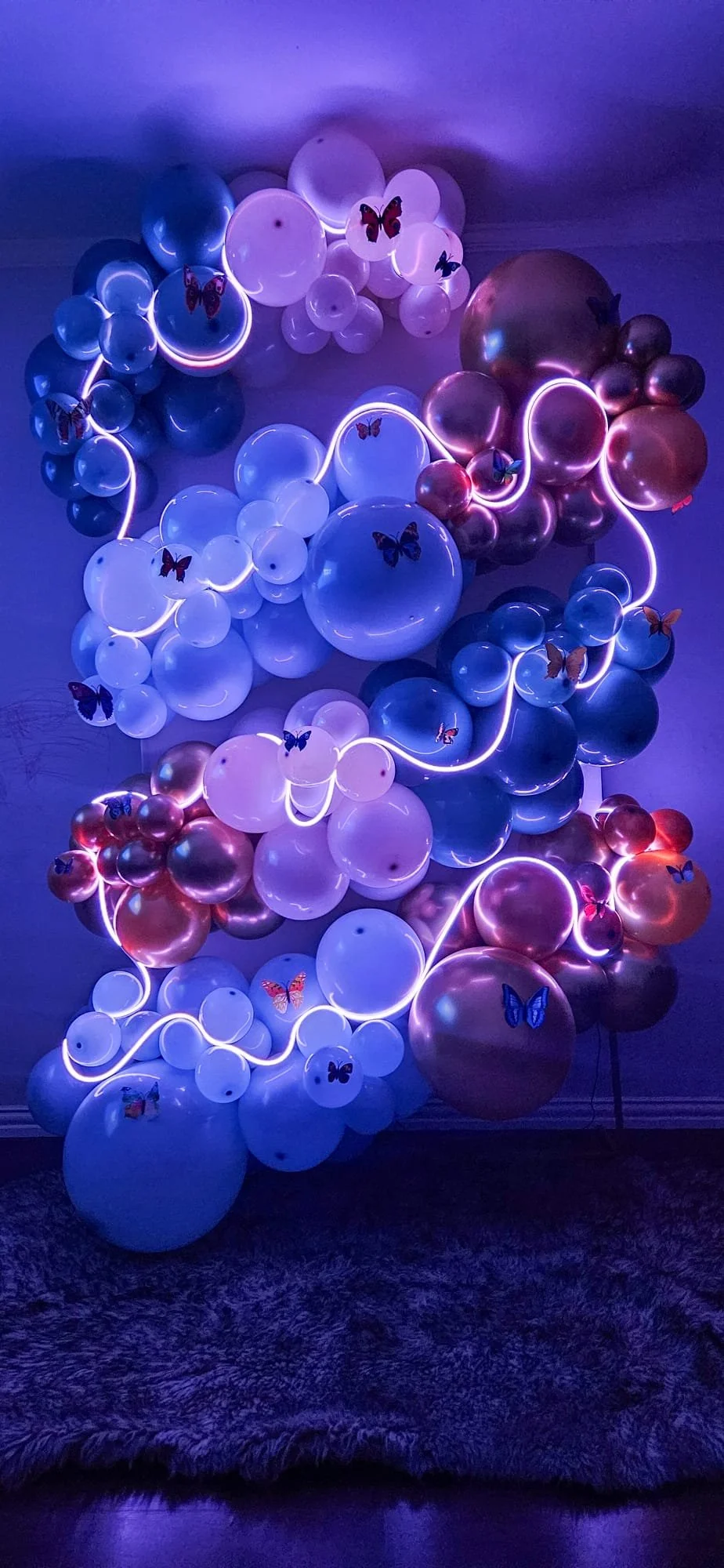 A decorative wall sculpture made of balloons in shades of white, pink, and purple, illuminated with purple and pink LED lights, adorned with colorful butterfly stickers.