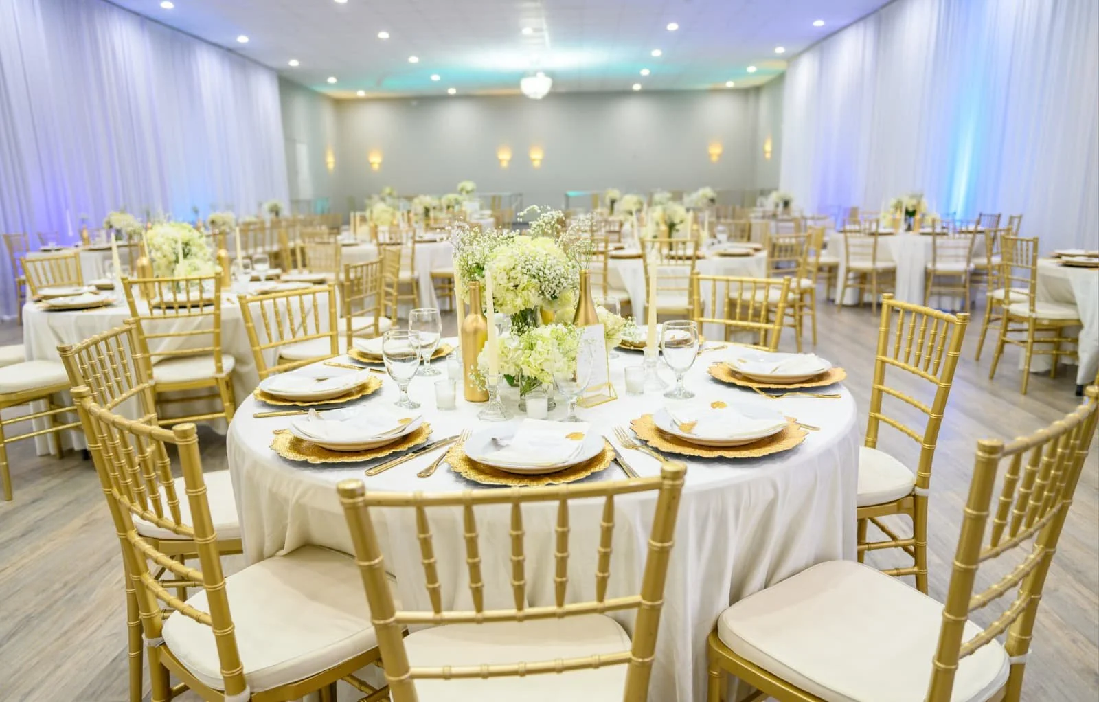 Elegant banquet hall decorated for a wedding or special event with gold chairs, white tablecloths, floral centerpieces, and gold accents.