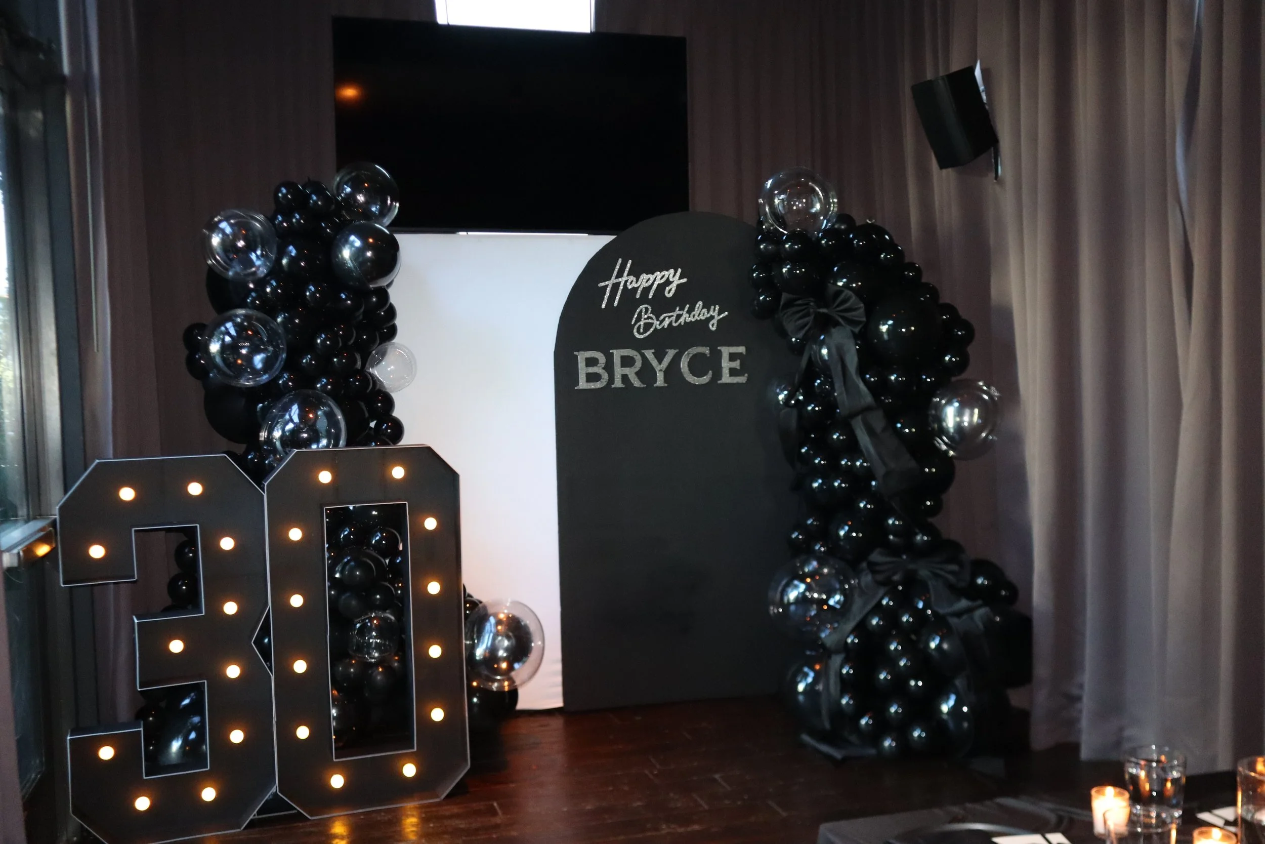 Black and silver balloon display with a black sign that reads 'Happy Birthday Bryce' in a decorated celebration area.